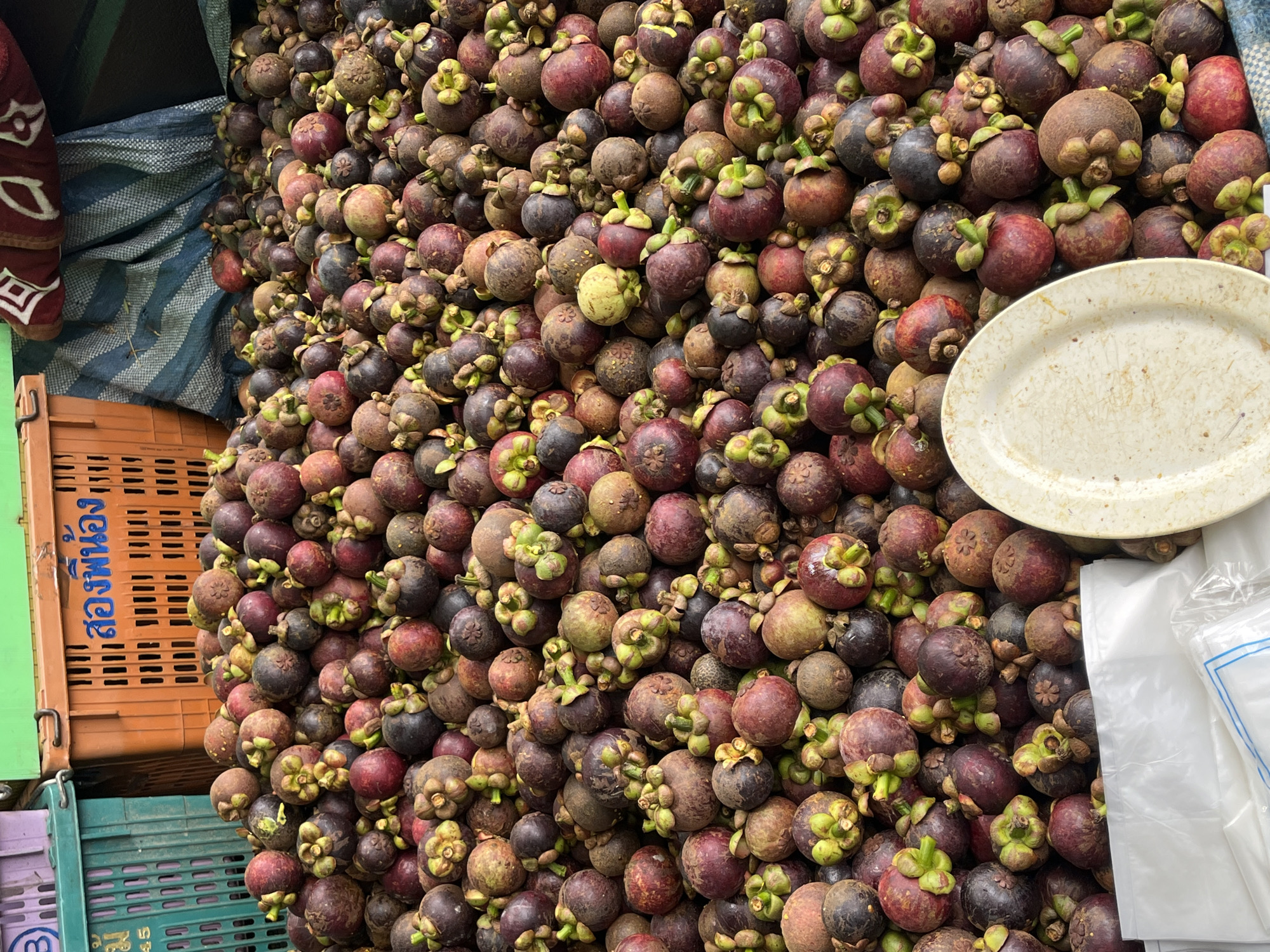 A large pile of mangosteens is displayed outdoors, likely at a market stall in Rawai, Thailand. The fruits, varying in shades of purple and brown, are densely packed and spill over the sides of orange and green plastic crates. A dirty, off-white plate rests on top of some of the mangosteens, partially obscuring the fruits beneath. White plastic bags are visible at the bottom of the frame. The background includes more crates and a striped blue and white tarp. The scene is brightly lit, suggesting it is daytime.