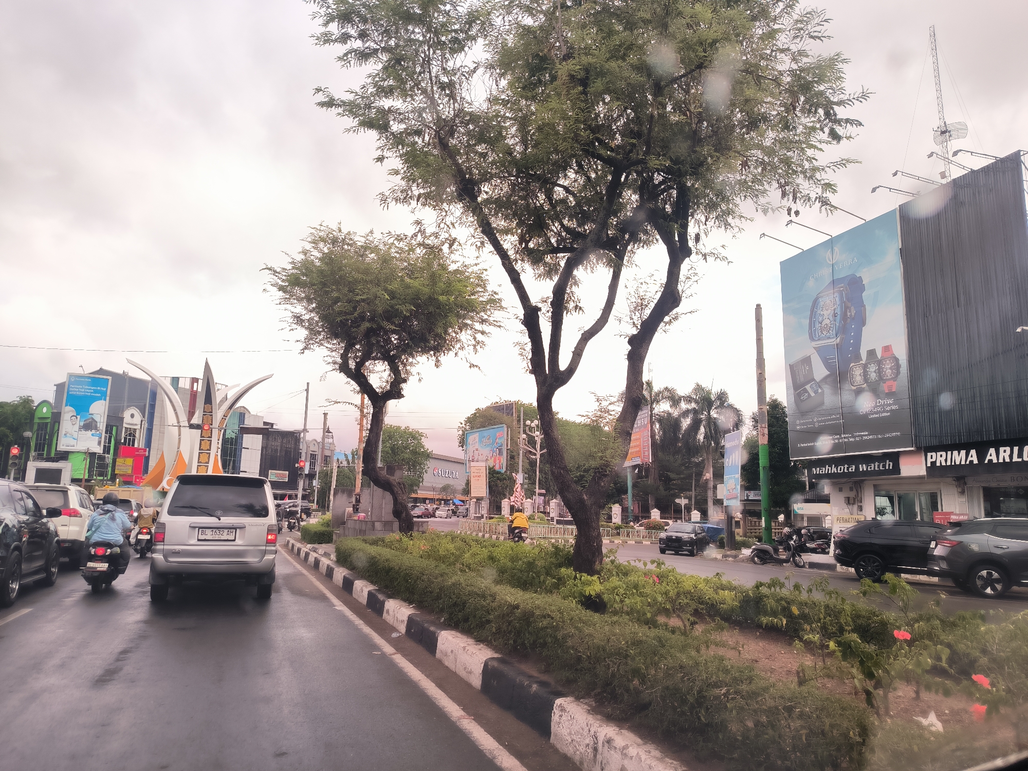 The photograph, taken through a wet windshield, captures a street scene in Banda Aceh, Indonesia. Traffic is visible, including cars and motorcycles. A silver minivan with license plate "BL 1632 AH" is a central subject. The road is wet, suggesting recent rain. Sidewalks are lined with black and white curbs. A median strip runs along the center of the road, planted with greenery. Various storefronts and billboards are present, including advertisements for "CHRIST VERRA" watches, "SUZUYA," and businesses named "Mahkota watch" and "PRIMA ARLO". Trees are positioned along the median and sidewalks. The sky is overcast. The photo suggests a typical day in a commercial area of the city.