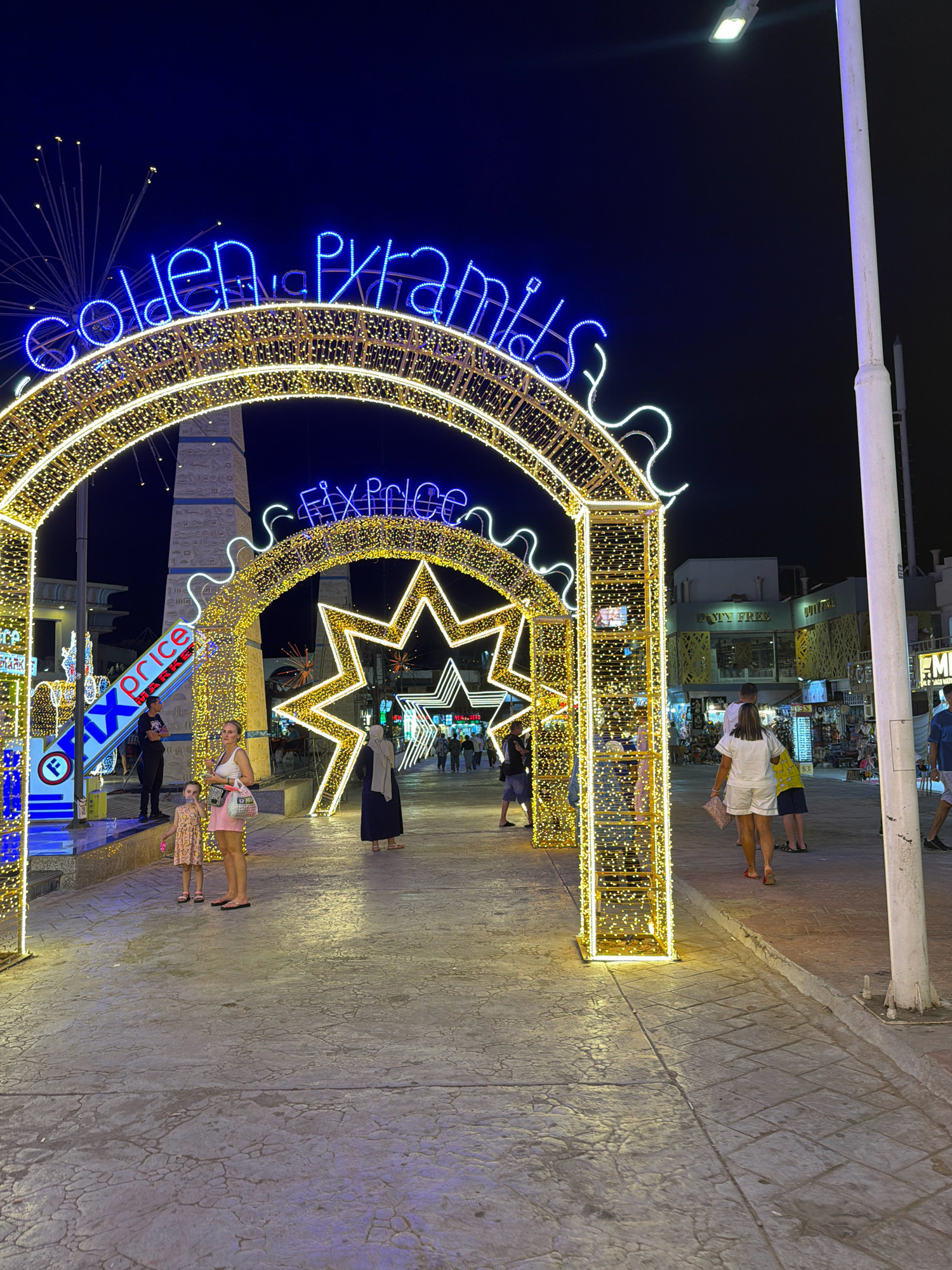 The image shows a nighttime scene in Sharm El Sheikh, Egypt, featuring decorative illuminated arches and stars. The arches are adorned with yellow lights, and a blue neon sign above reads "GOLDEN PYRAMIDS." Another blue neon sign, partially visible, says "FIX PRICE." People are walking along a paved area under the arches. In the foreground, a woman and a young girl are standing, with the woman looking towards the camera. Behind them, other individuals are seen walking and browsing. Shops with illuminated signs are visible in the background, including one labeled "DUTY FREE." The environment appears to be an outdoor pedestrian area, likely a promenade or marketplace, decorated for an evening event or attraction.