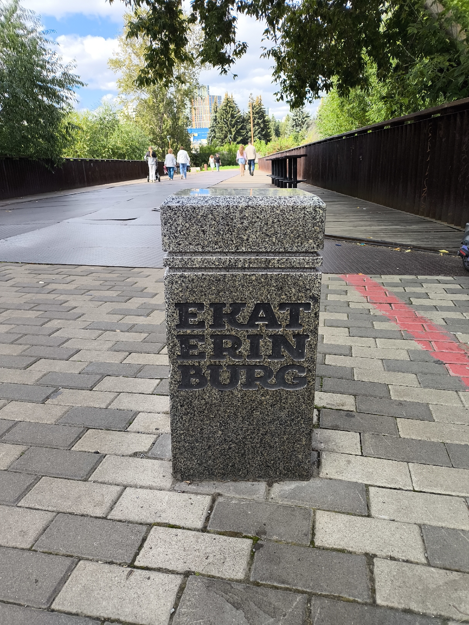 In Yekaterinburg, Russia, on a paved walkway, a granite pillar stands with the inscription "EKATERINBURG" carved into its surface. The pillar is positioned on a path made of grey and beige rectangular pavers. To the right of the pillar, a red stripe is painted across the pavers, forming a diagonal line. Beyond the pillar and walkway, a wooden bridge or elevated path is visible, bordered by a dark brown metal railing. Trees with green foliage line the sides of the path, and in the background, modern buildings with windows are visible. Several people are walking on the elevated path, with some pushing strollers. The sky above is blue with scattered white clouds.