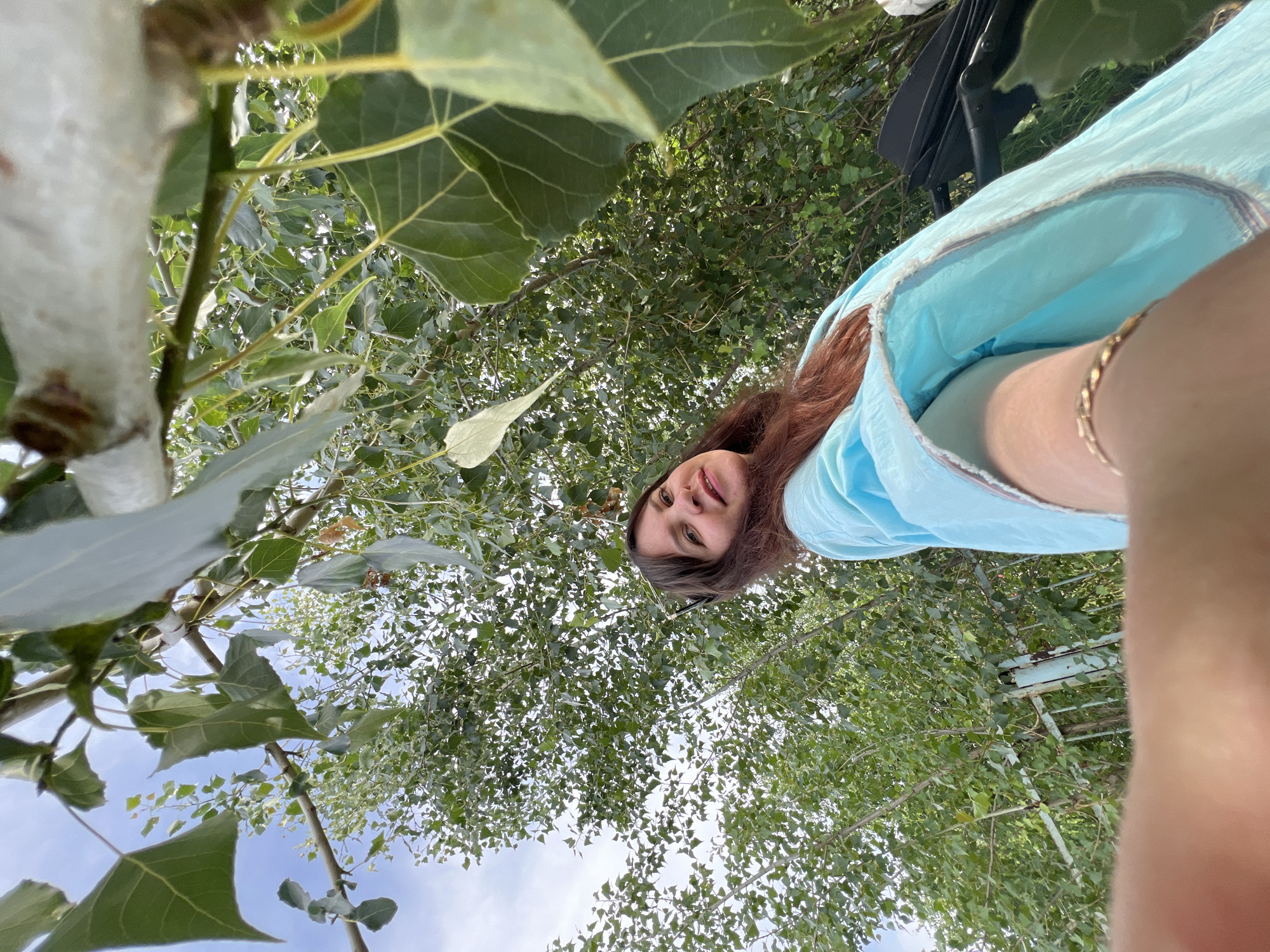 The image captures a woman in a bright blue outfit posing for a selfie under a tree. She has long, reddish-brown hair and is smiling. The background is filled with lush green leaves and branches of a tree, with a glimpse of a bright blue sky. A weathered metal fence is visible behind her, suggesting an outdoor setting. A stroller is partially visible to her right. The perspective is from a low angle, looking up towards the woman and the tree canopy. The presence of the stroller hints at the woman possibly being a parent. The location is identified as Noviy, Russia.