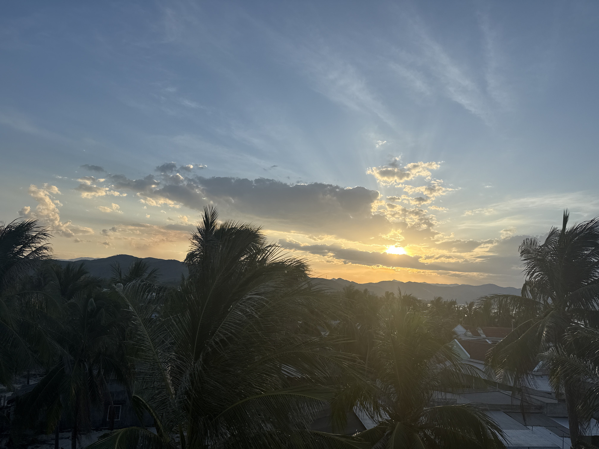 The image captures a sunset over a tropical landscape. In the foreground, lush palm trees with their fronds dominate the scene, creating a natural frame. Beyond the trees, a cluster of buildings with red-tiled roofs can be seen, suggesting a residential area or small town. In the distance, rolling hills or mountains form a dark silhouette against the colorful sky. The sky itself is a spectacle of light and clouds, with the sun just beginning to dip below the horizon, casting a warm, golden glow. Rays of sunlight pierce through the scattered clouds, creating a dramatic and serene atmosphere. The overall scene conveys a sense of tropical tranquility and the beauty of a sunset in an undisclosed location.