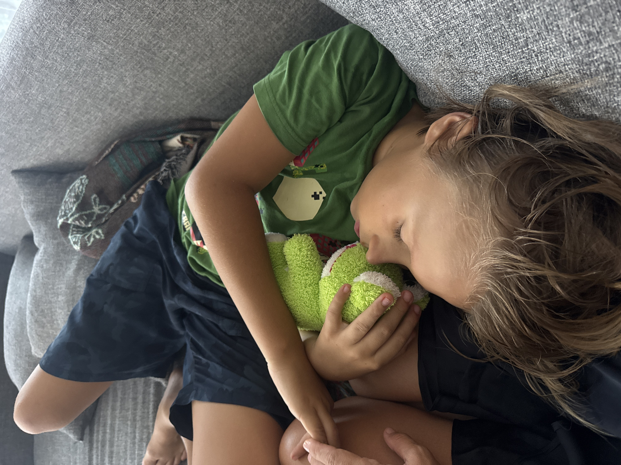 An overhead shot captures a young child soundly sleeping or resting on a grey upholstered sofa, cradled by an adult who is partially visible. The child, with light brown, tousled hair, is wearing a green short-sleeved t-shirt featuring a pixelated white figure (resembling a Minecraft Ghast) and red accents, paired with dark blue patterned shorts. The child’s eyes are closed, and lips slightly parted, conveying a deep state of repose. Clutched gently in their hands is a bright green, fluffy stuffed toy with white trim, which the child holds close to their face. An adult's hands are visible, providing comfort by gently holding the child's leg/thigh.
The setting appears to be indoors, likely a living room, in a calm and peaceful atmosphere. A patterned blanket or throw is partially visible near the child's feet, tucked into the sofa. The overall scene suggests a quiet moment of rest or a nap. The location context is Kuta Selatan, Indonesia. No specific time of day or weather is discernible, but the lighting appears natural and soft. There is no legible text visible on the child's shirt, only the pixelated graphic design.