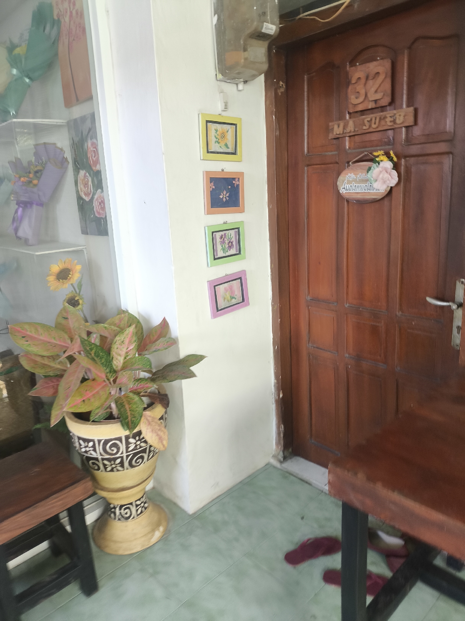 The image depicts a close-up view of a wooden door with the house number "32" and the name "M.A. SU'ED" above a decorative sign that reads "Assalamualaikum." The door is flanked by a cream-colored wall adorned with four small framed pictures of floral motifs. To the left of the door, a potted plant with broad, variegated leaves sits next to a dark wooden stool. Further left, a glass display case showcases bouquets of flowers and artwork. The floor is a light green tiled surface, and a burgundy flip-flop is visible near the bottom right corner of the frame. The scene appears to be an entrance to a residence in Lumajang, Indonesia, possibly a shop or a home.