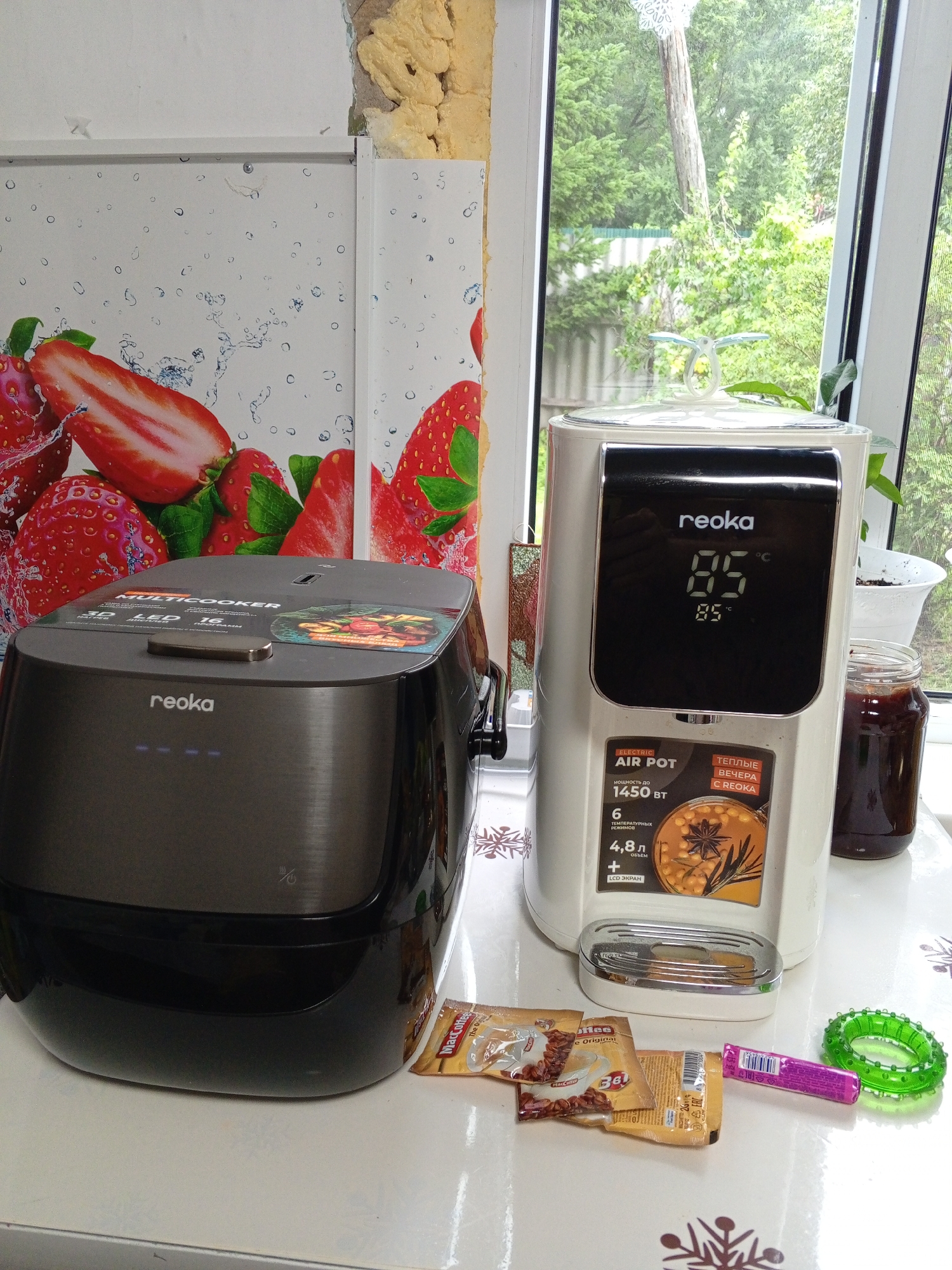 The image shows a domestic scene featuring two kitchen appliances, a multicooker and an electric air pot, placed on a white tabletop. The multicooker, on the left, is dark gray with a brushed metal finish and a black base. It has the brand name "reoka" printed on it. The electric air pot, on the right, is white and also has the "reoka" logo. Its digital display shows "85 C". Next to the air pot is a glass jar containing a dark substance, possibly jam or preserves. In front of the appliances, there are a few small packets of instant coffee and a green ring-shaped object, possibly a toy or decoration. The background includes a window with a view of greenery outside and a wall decorated with a strawberry pattern. The lighting suggests it is daytime. The scene appears to be indoors, likely a kitchen.