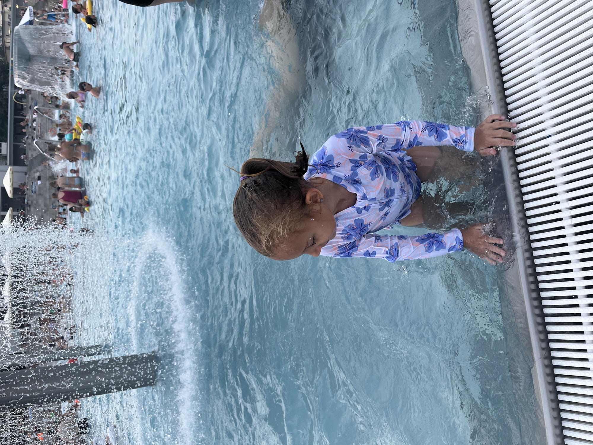 In this image, a young girl with brown hair is in a swimming pool. She is wearing a long-sleeved rash guard with a blue and white floral pattern and her arm is resting on the edge of the pool, which has a white grate. The girl is looking down into the water. The pool appears to be crowded with other people swimming and playing. The water is a clear, bright blue. In the background, there is a structure with a waterfall or fountain cascading down, creating a misty effect. The overall scene suggests a sunny day at a public pool or water park.