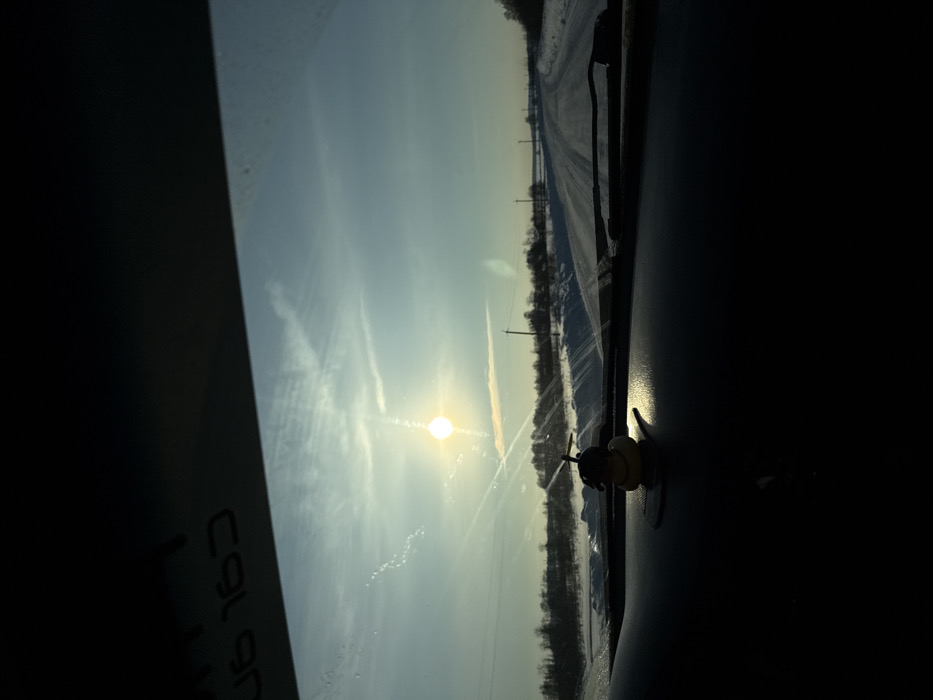 The image captures a sunlit, snowy winter landscape viewed from inside a vehicle on a rural road near Dubrova, Russia. The low sun, a brilliant white orb with a starburst effect, is positioned in the upper left, casting strong light across the scene and creating glare on the windshield. The sky is a pale blue with faint contrails and high cirrus clouds. A two-lane road, covered in snow with visible vehicle tracks, extends into the distance, flanked by snow banks and bare trees. Several utility poles with power lines are visible along the right side of the road. The dark dashboard of the vehicle occupies the lower portion of the frame, featuring a small, yellow, duck-shaped ornament wearing a propeller helmet, illuminated by the sun. Reflected text, appearing as "CGL 90" or "C9L 90" in reverse, is visible on the upper left section of the windshield. No individuals are present.