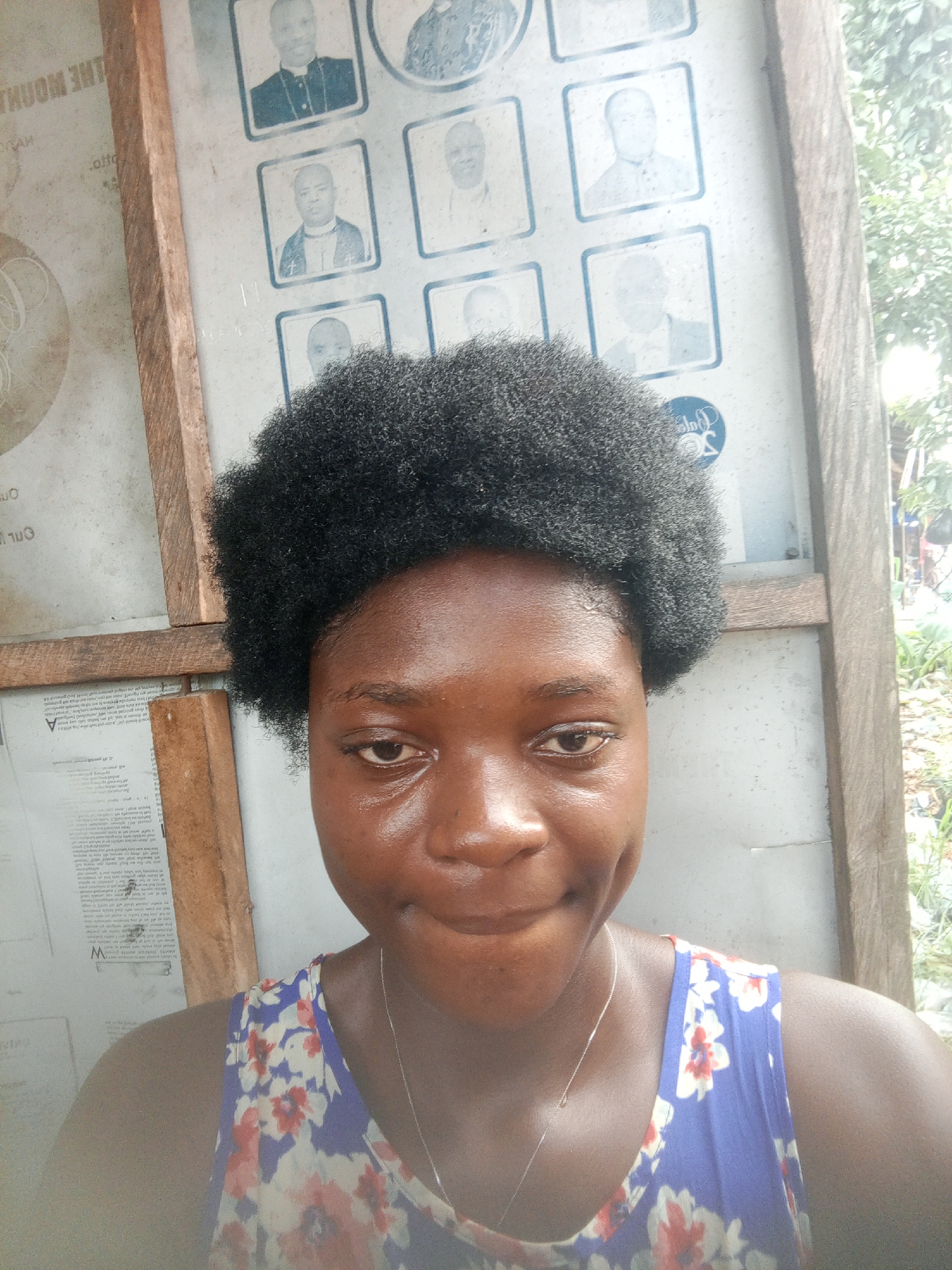 A young, dark-skinned woman is the main subject of the photograph. Her black, short, curly hair frames her face. She has a dimple on her left cheek and is wearing a silver necklace. Her attire consists of a sleeveless top with a floral pattern on a blue background. She is positioned in front of a weathered, wooden-framed board, which displays several portrait images of men in clergy attire. Attached to the left side of the board are papers with writing. The photo appears to be taken outside in the city of Mbribit, Nigeria, evident by the foliage and environment in the background on the right side of the board.