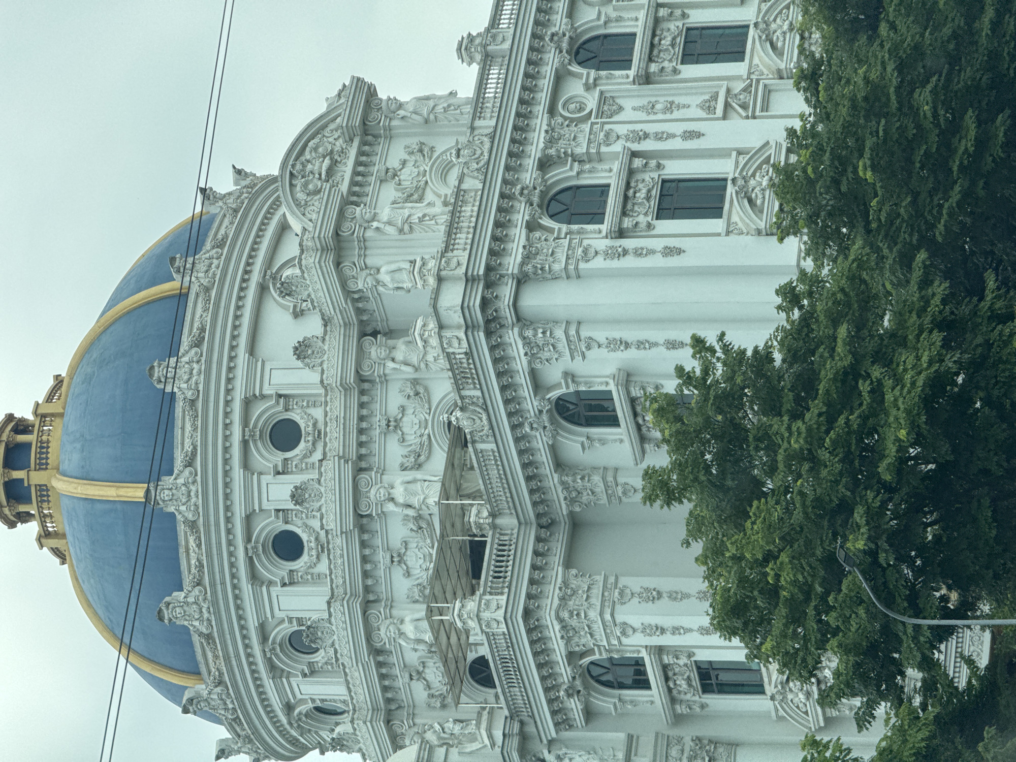 The image shows a portion of an ornate white building with a blue and gold dome, located in Hoa Lư Ward, Vietnam. The building's facade is decorated with intricate carvings, statues, and arched windows. There are trees in the foreground, and power lines are visible against the overcast sky. No people are visible in the image.