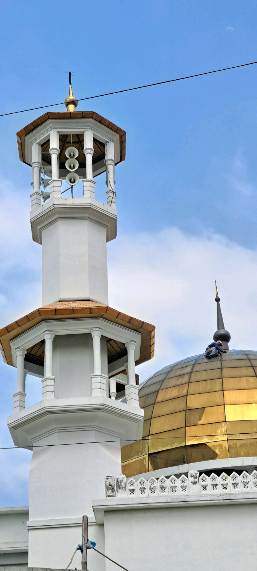 This is a low-angle, vertical, eye-level shot of the minaret and dome of a mosque in Petungasri, Indonesia. The sky is visible in the background with a few scattered clouds. The minaret is white with a gold-colored roof and a gold finial. It has an octagonal shape and features three white speakers mounted on the upper section. The dome is large, golden, and has a pointed black finial. A person wearing a blue shirt and a white hard hat is visible on top of the dome, appearing to be working. A decorative white railing with intricate designs is visible on the lower section of the mosque. Wires are also present in the foreground, and a thin black wire runs horizontally across the image.