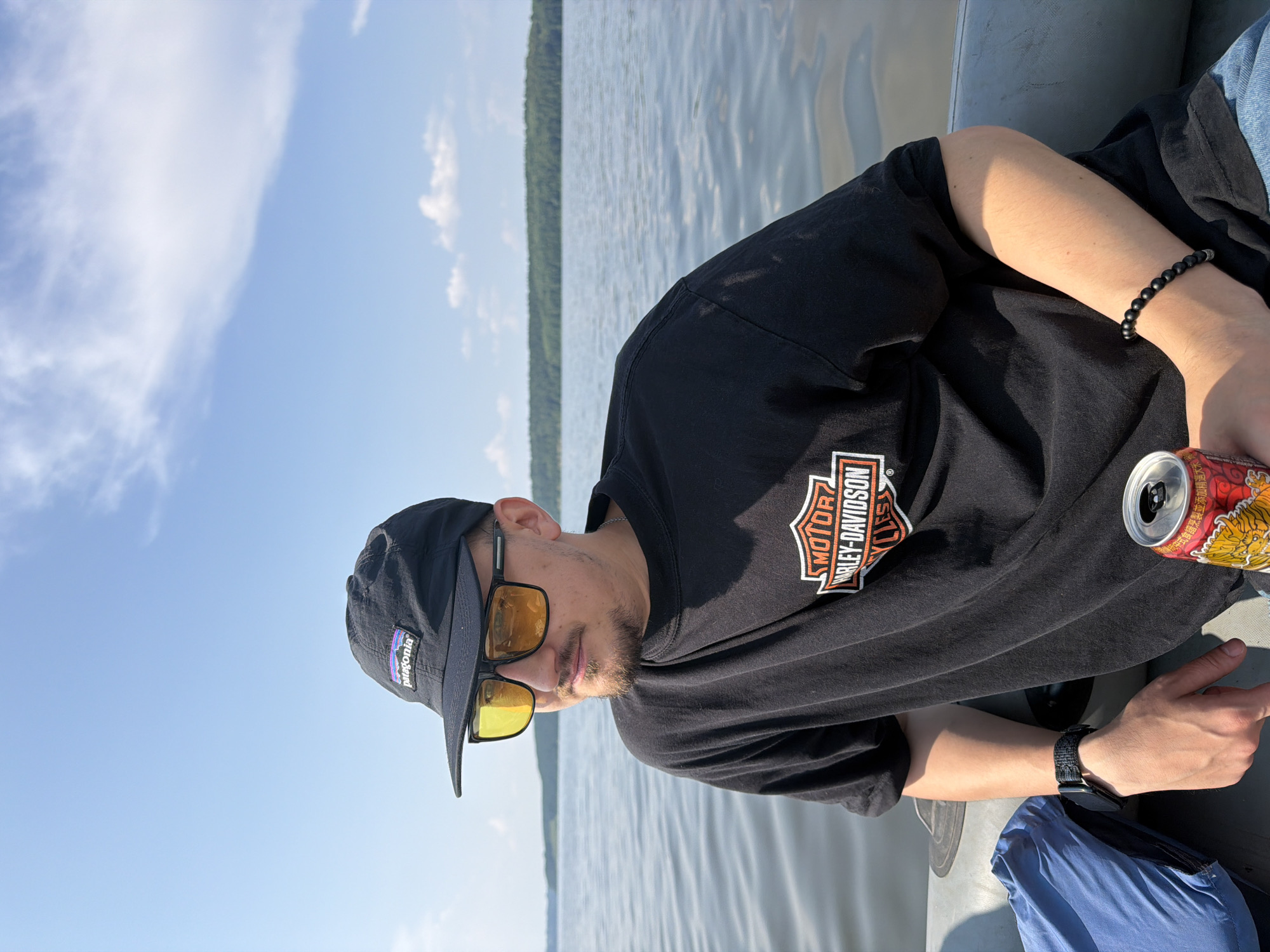 A young man is sitting on an inflatable boat in a lake. He is wearing a black baseball cap with a Patagonia logo, yellow-tinted sunglasses, and a black t-shirt with a Harley-Davidson logo. He has a light beard and is looking to his left. In his right hand, he is holding a red and gold can of what appears to be a beverage. He is also wearing a black watch on his left wrist and a beaded bracelet on his right wrist. The boat is grey and a blue bag is visible on the left side of the frame. The background shows a calm lake with trees and a blue sky with scattered clouds. This scene does not appear to be in Sylva, Russia.