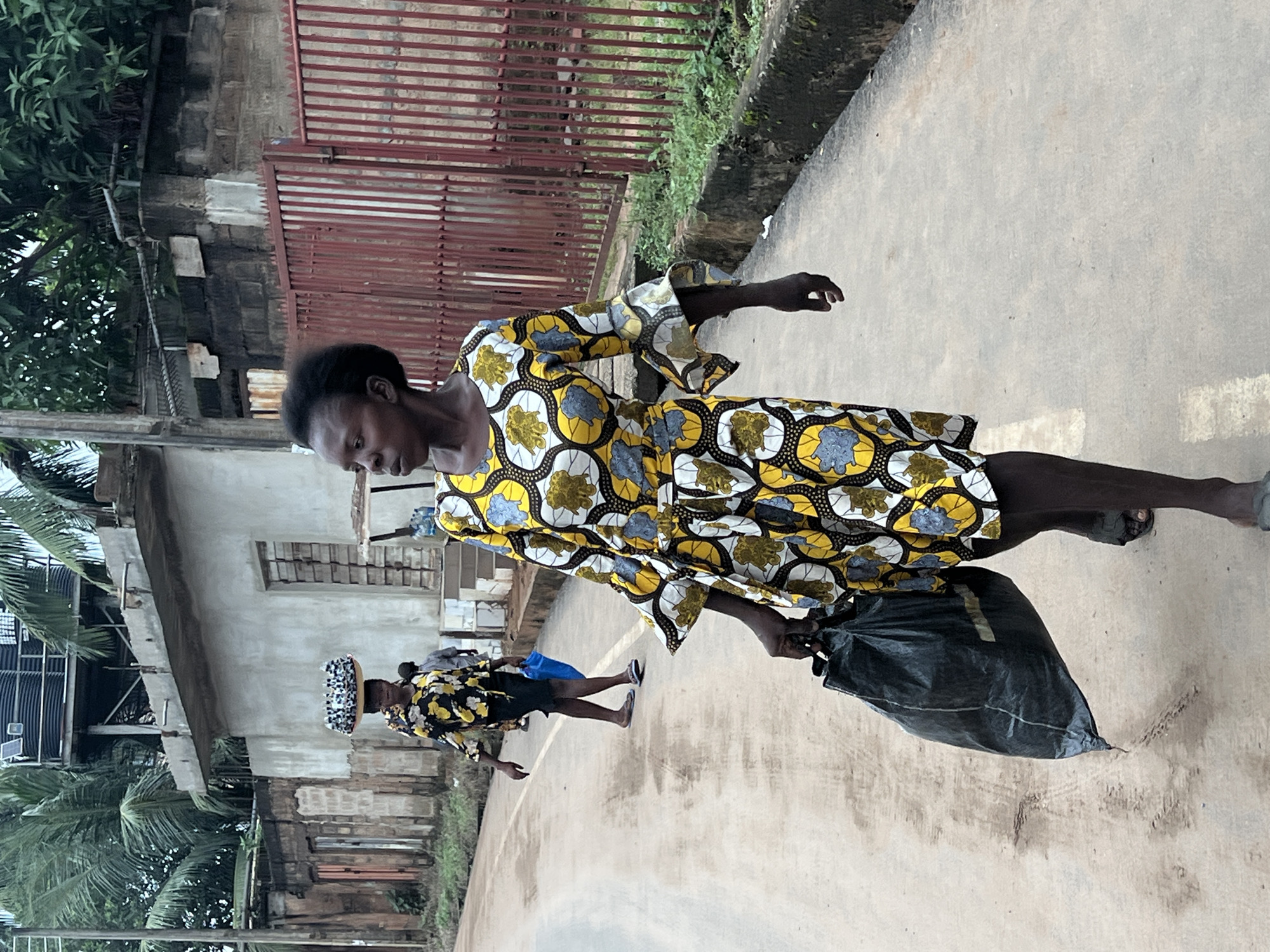 In Nkpor, Nigeria, on a paved road, two women walk. The woman in the foreground wears a brightly colored dress with a yellow, white, and blue floral pattern and carries a large black plastic bag in her right hand. She walks towards the right side of the frame. Behind her and to the left, a second woman walks away from the camera, carrying a tray of items on her head, and a blue plastic bag in her left hand. The background features buildings with rough plaster walls, a red metal fence, and lush green palm trees and other foliage. The ground is a light brown color with some tire tracks and debris visible.