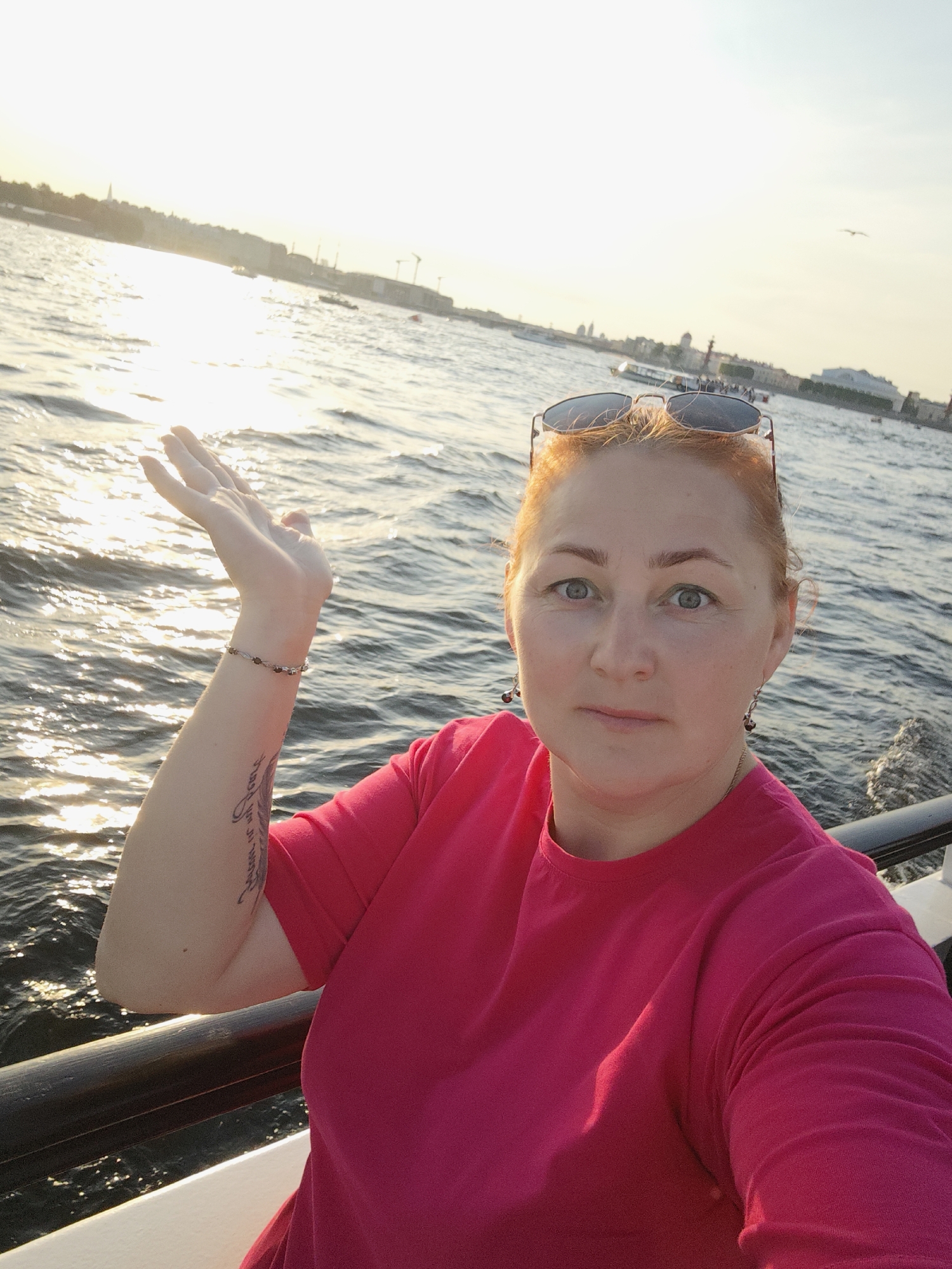 In Saint Petersburg, Russia, a woman with fair skin and red hair, wearing a pink t-shirt and sunglasses perched on her head, poses for a selfie from a boat. The sun is setting over the water, creating a bright glare. In the background, a city skyline with buildings and cranes is visible. The woman has a tattoo on her left arm and is wearing earrings and a bracelet. She is holding her left hand up, palm facing outwards, towards the sun. The boat has a dark railing in the foreground.