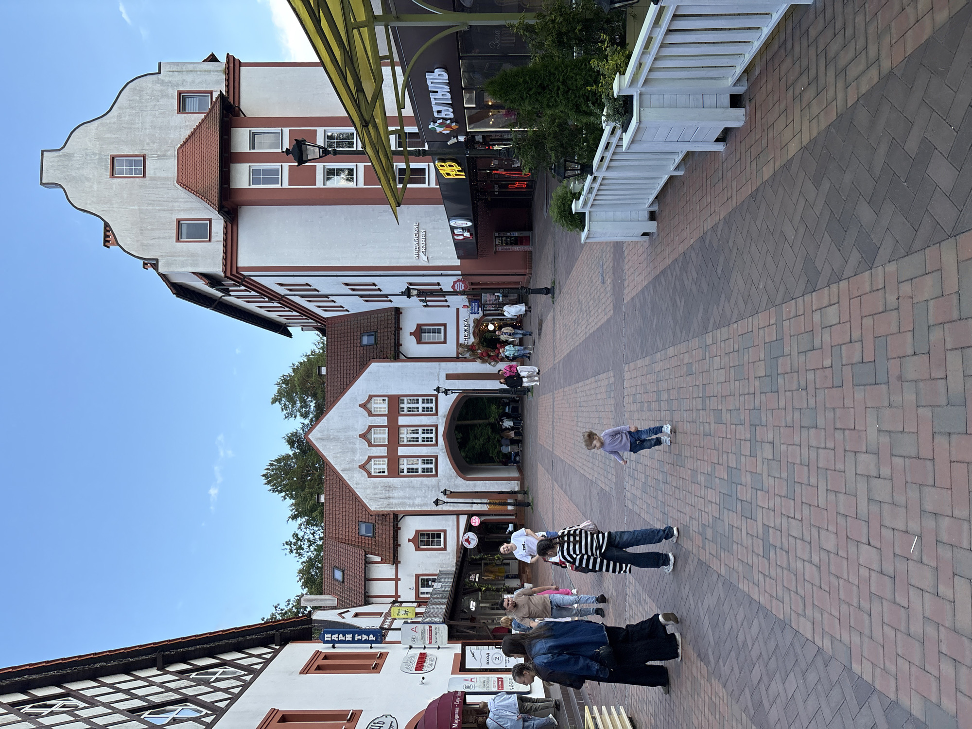 A wide cobblestone walkway is lined with buildings in a European architectural style. On the left, a white building with dark timber framing and red trim features multiple windows. Further down, a white building with a red roof has an archway leading to a darker area. On the right, a tall, multi-story building with red and white accents dominates the background. A yellow awning extends over shops on the ground floor of this building, one of which has a prominent sign with Cyrillic lettering. Several people are walking on the path. In the foreground, a young child in a purple shirt and jeans walks away from the camera towards the right. Two women and a person in a denim jacket are also in the foreground on the left. A woman in a black shirt and black pants is to their right, and a child in a striped shirt and jeans is in the middle. The sky is a clear blue with a few scattered clouds. The environment appears to be a pedestrian zone, possibly a tourist area in Svetlogorsk, Russia.