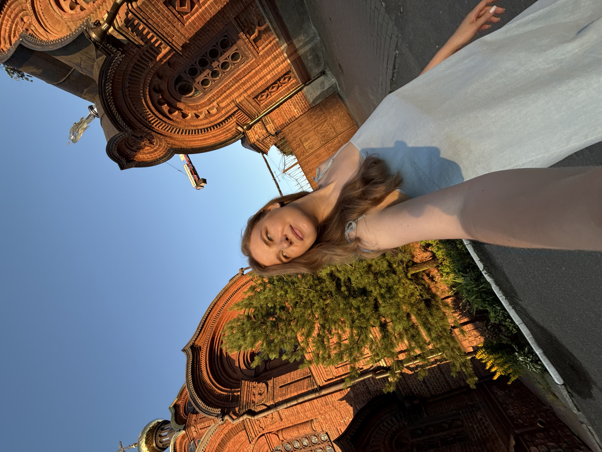 A young woman with long, light brown hair is taking a selfie in front of a red brick church. She is wearing a light blue dress. The church has golden domes and intricate architectural details. A green pine tree is visible to the left of the woman, and a decorative metal gate is to the right. The sky is clear and blue, with the warm glow of sunset illuminating the scene. This is likely the Assumption Cathedral (Uspensky Sobor) in Vladimir, Russia, known for its striking red brick architecture and golden domes.