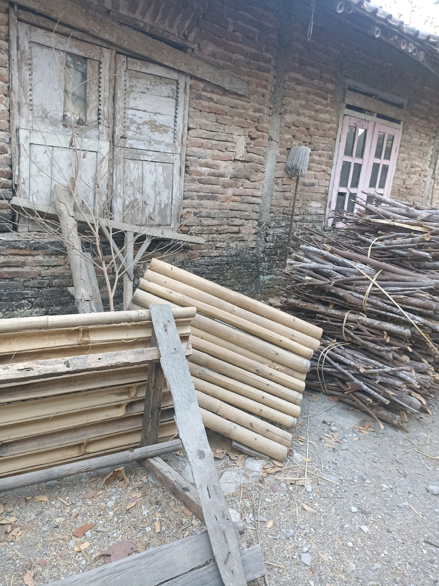 A weathered brick building with a tiled roof stands in the background. In front of the building, a pile of cut branches and logs is stacked to the right. To the left of the branches, a section of bamboo poles is neatly arranged. A wooden door with peeling white paint and a small window is visible on the left side of the building, partially obscured by a bare branch. On the right side of the building, a pink-framed window can be seen. A mop with a white head on a metal pole leans against the wall near the center of the image. The ground in front of the building is covered with gravel and dry leaves. The scene appears to be an outdoor area in Widoro, Indonesia, possibly a backyard or a storage area for building materials.