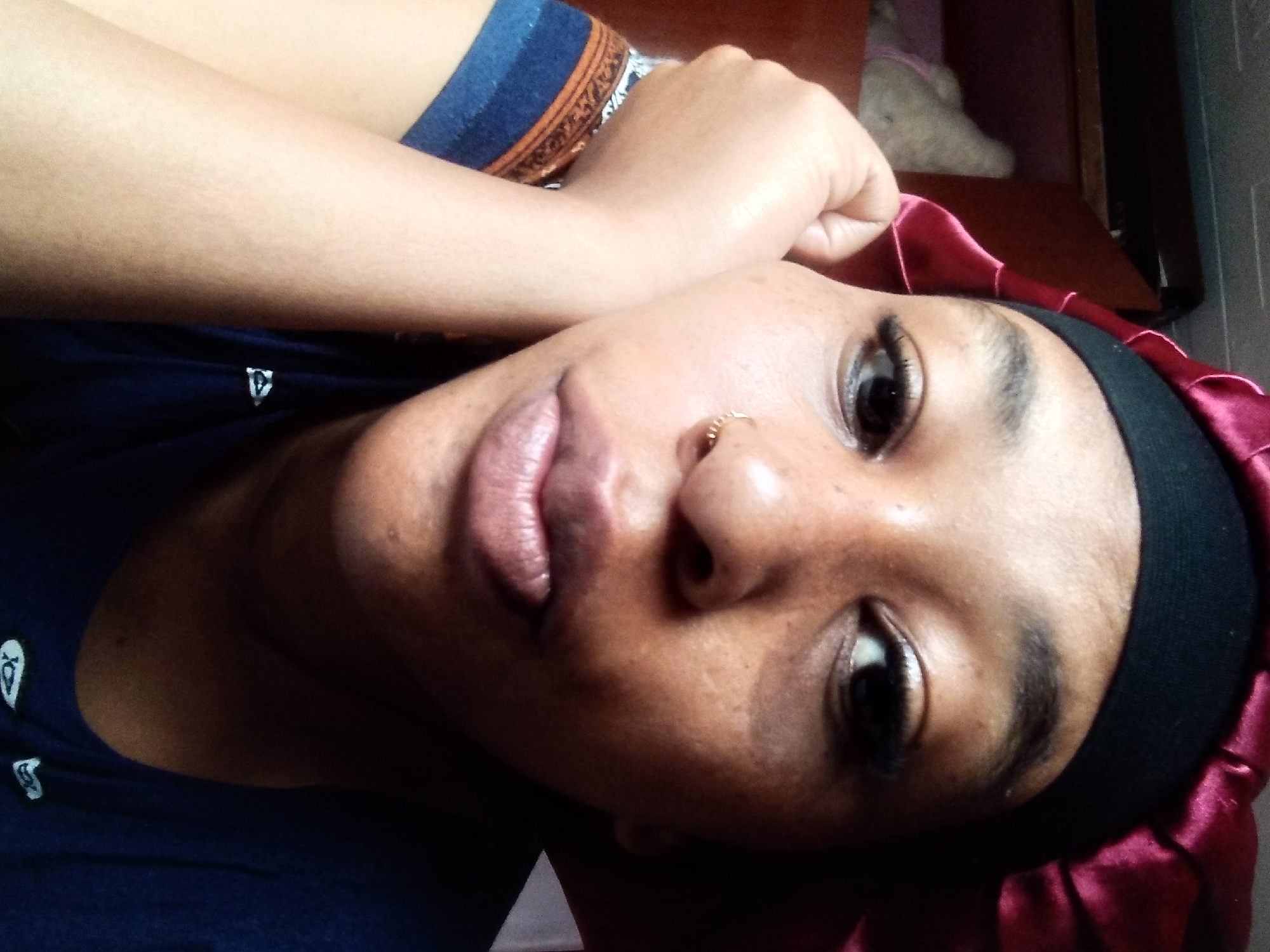 A woman is pictured from the chest up, indoors, with her head tilted. She is wearing a dark blue t-shirt and a burgundy satin bonnet. Her right arm is bent and rests against her face. She has a small gold nose ring and what appear to be false eyelashes. In the background, a shelf with a stuffed animal is visible. The image is a close-up, personal photograph, likely taken by the subject herself. There is no indication of a specific location in Nyeri, Kenya.