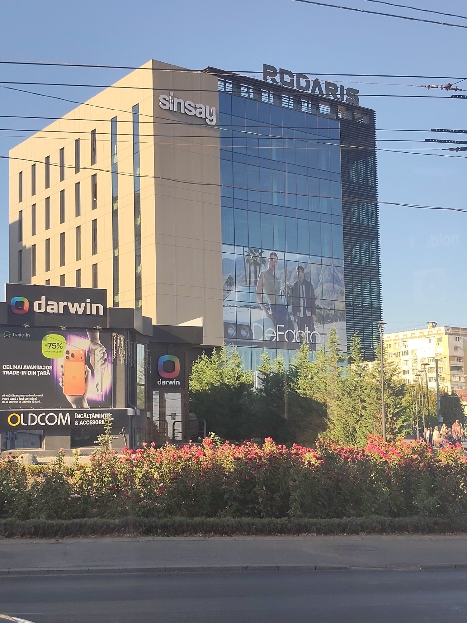 The image shows a modern office building in Chișinău, Moldova. The building has a light-colored facade on the left and a glass-covered section on the right. The "Sinsay" logo is visible on the light-colored section, and the "Rodaris" logo is at the top of the glass section. A large advertisement for "DeFacto" featuring two men and a car is displayed on the glass facade. Below the main building, there are storefronts with "Darwin" logos. One storefront has a billboard advertising a trade-in offer for mobile phones, showing an orange smartphone and text in Romanian. Another sign below that advertises "OLDCOM" and "ÎNCĂLTĂMINTE & ACCESORII" (Footwear & Accessories). A row of red roses and green bushes lines the sidewalk in front of the stores, with trees and other buildings visible in the background. The sky is clear and blue. The scene appears to be captured during the daytime.