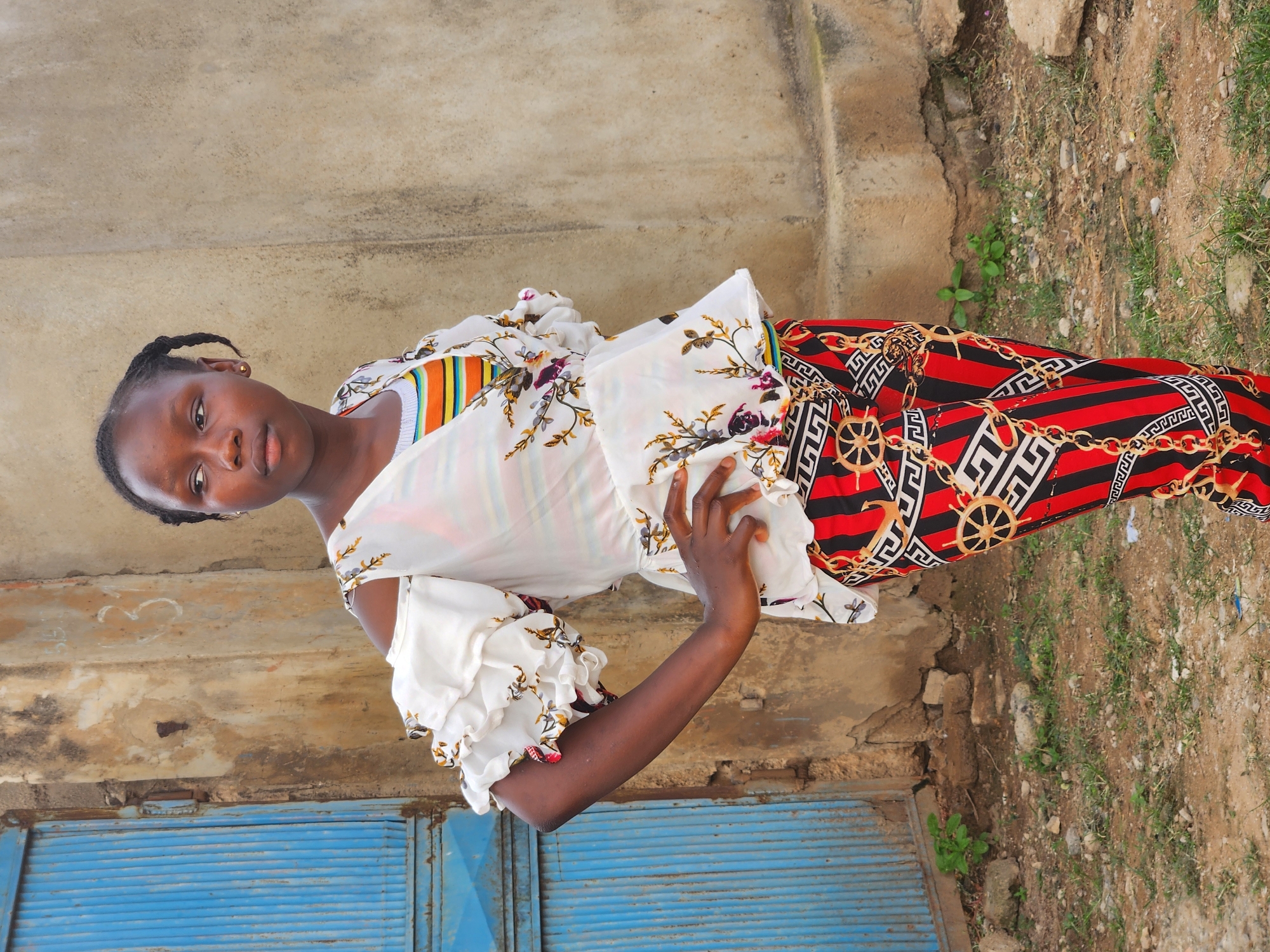 In Sabon Tasha, Nigeria, a young Black female stands outdoors, posing for a photo. She is positioned slightly off-center to the right, with her body angled towards the left. The background consists of a textured, weathered wall and a blue metal door. The ground is dirt with scattered small rocks and patches of green grass.
The female wears a white off-the-shoulder blouse with ruffled sleeves, adorned with a floral pattern. Beneath this, she wears a striped tank top in orange, yellow, green, and white. Her pants are brightly patterned with vertical red and black stripes, interwoven with a gold chain and Greek key design. Her hair is styled in two braids that fall over her shoulders. She has a gold earring in her visible ear. Her left hand rests on her hip, and her right arm is bent, with her hand near her waist. She looks to her left, away from the camera.