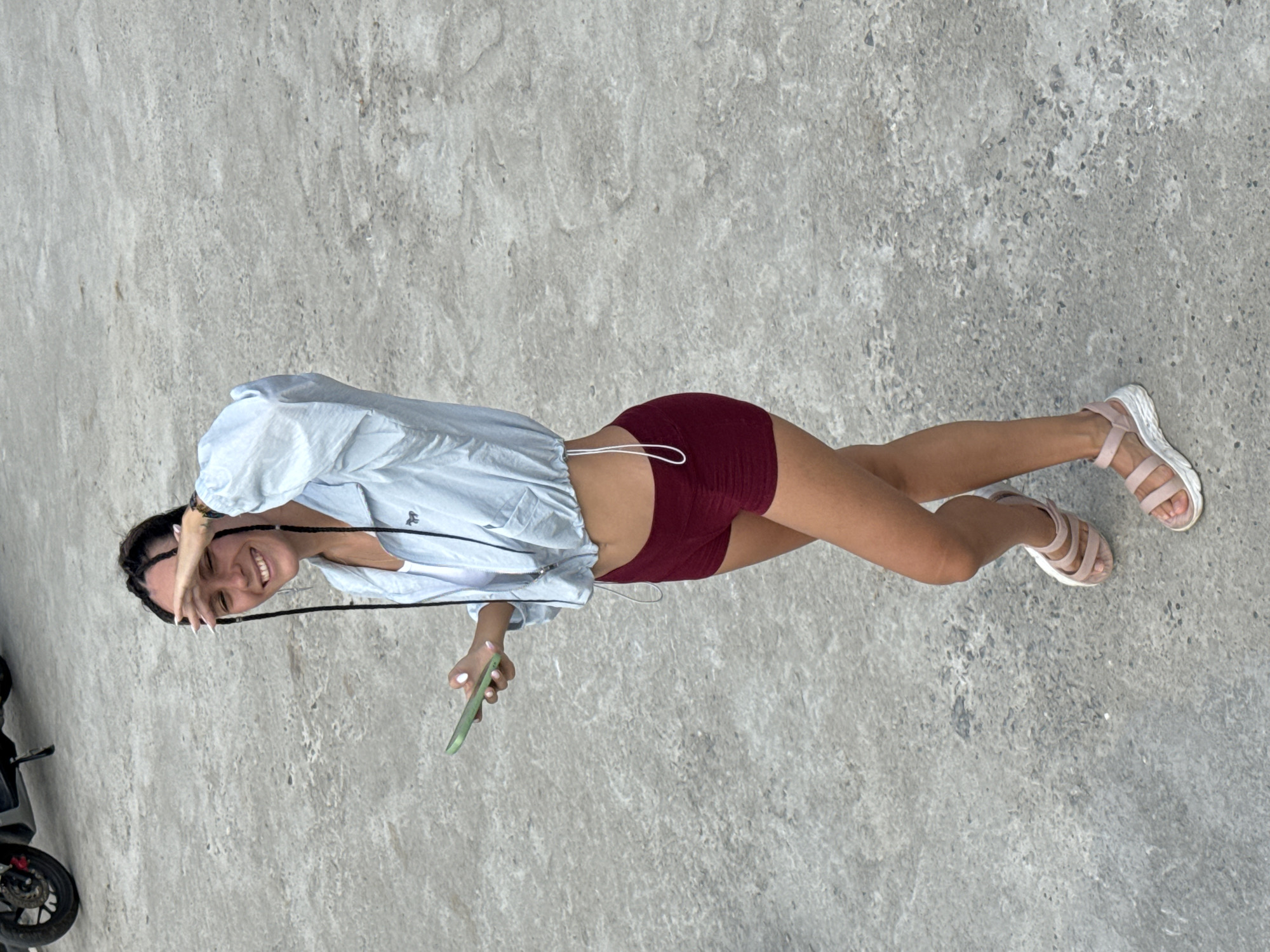 A young woman is standing on a concrete surface outdoors. She is wearing a light blue long-sleeved shirt, red shorts, and pink sandals. She is holding a green smartphone in her right hand and obscuring her face with her left hand. She is smiling. There is a black motorcycle partially visible on the left side of the image. The location is presumed to be somewhere within Lũng Phìn Commune, Vietnam, as specified in the request. The background consists of a textured concrete surface.