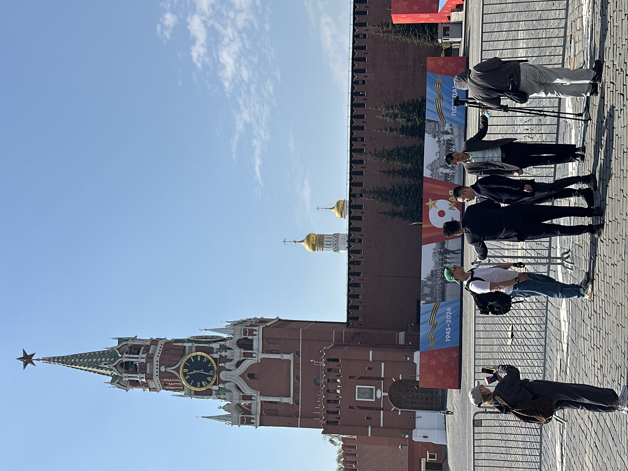 The image shows a street scene in Moscow, Russia, featuring the Kremlin's Spasskaya Tower and part of the Kremlin wall. The clock on the Spasskaya Tower reads approximately 01:52 PM. The sky is clear with some clouds. Along the Kremlin wall, there are banners that appear to be related to a celebration or commemoration. A group of people is present, some standing near the banners and others taking pictures. Some people appear to be part of a media crew.