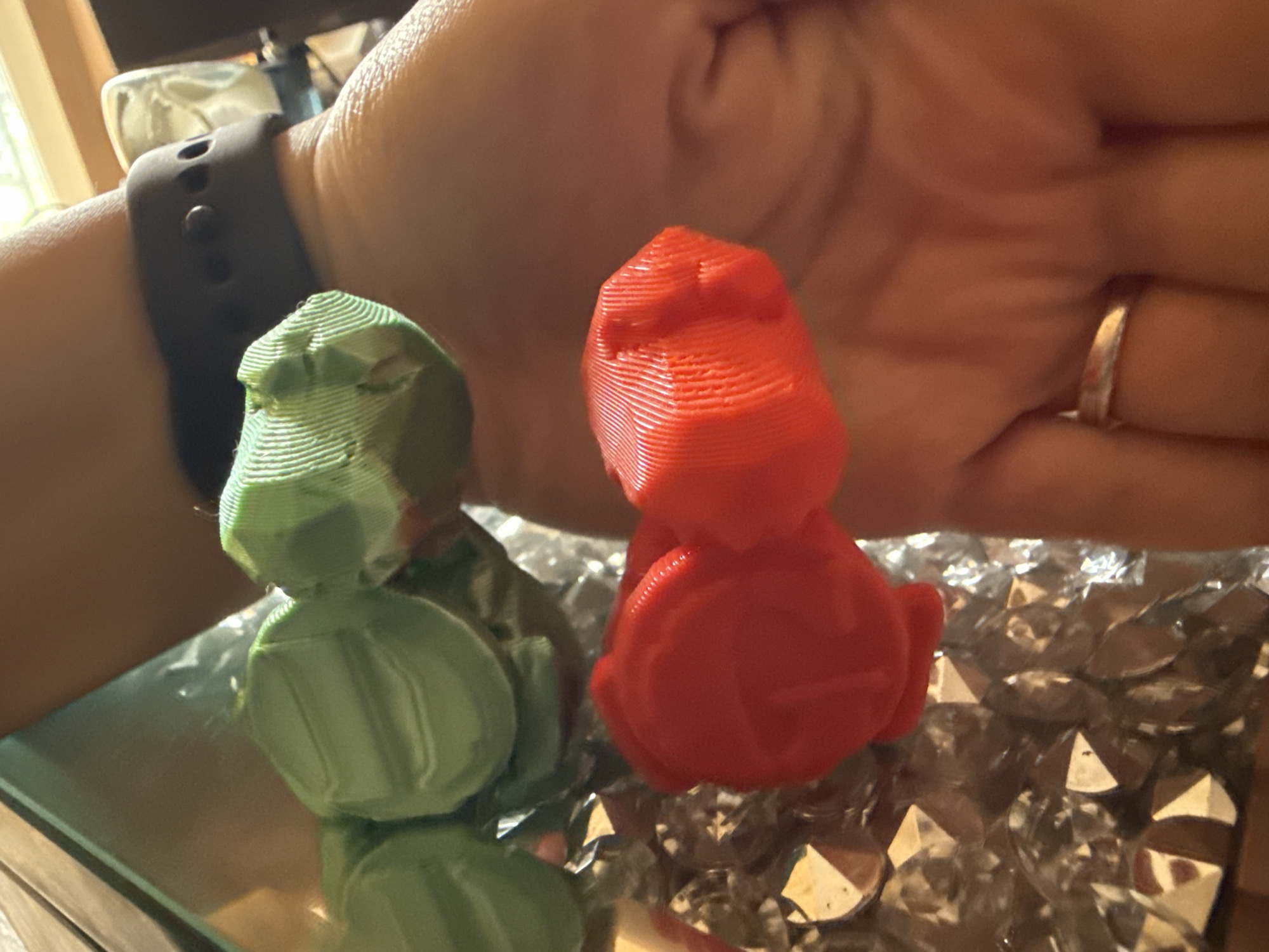 A close-up shot shows two small, 3D-printed figures, one green and one red, resting on a reflective surface covered in faux diamonds. A human arm with a black smartwatch on the wrist is visible in the background, with a hand and a ring on one of the fingers further back. The location and specific city remain unknown.