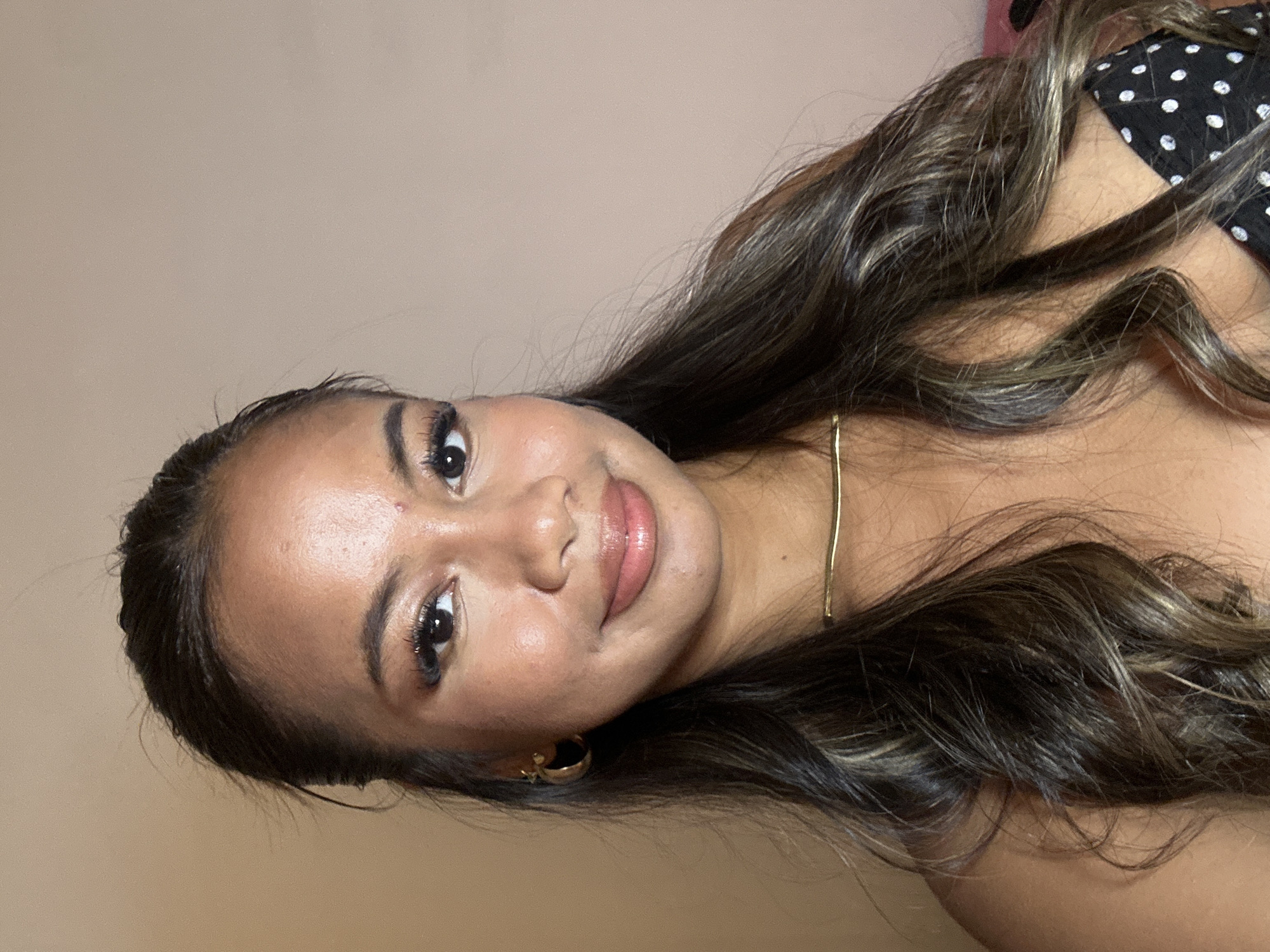 A woman with dark hair is facing the camera. She has light brown skin, dark eyes, and full lips. She is wearing a gold necklace and gold hoop earrings. She is wearing a black and white polka dot top. The background is a pale beige color. The image appears to be taken indoors. It is difficult to ascertain from the image whether or not this was taken in Celuk, Indonesia.