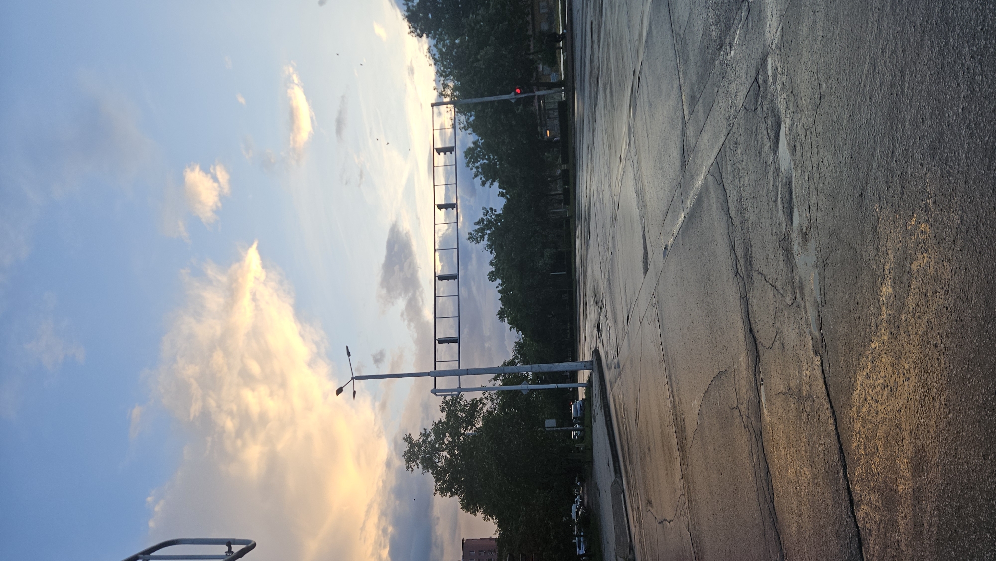 The image depicts a street scene, possibly in Plovdiv, Bulgaria, though without additional landmarks this cannot be verified. The foreground shows a cracked, asphalt road surface. A traffic light structure with signals is positioned vertically on the left, adjacent to trees and parked cars along the roadside. The sky above exhibits clouds and hints of sunset or sunrise colors. The scene appears to be photographed in a slanting angle.