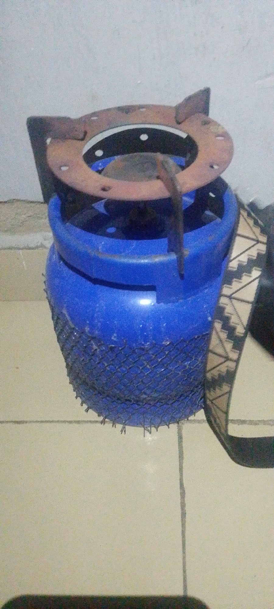 An improvised cooking stove, fashioned from a blue gas cylinder, is situated on a tiled floor in Jauro Yinu, Nigeria. A rusty metal grate, presumably for supporting cookware, is affixed to the top of the cylinder. The cylinder itself is partially wrapped in a dark mesh, held in place by what appears to be a patterned strap or belt. The background consists of a textured, light-colored wall. The lighting is dim, casting soft shadows.