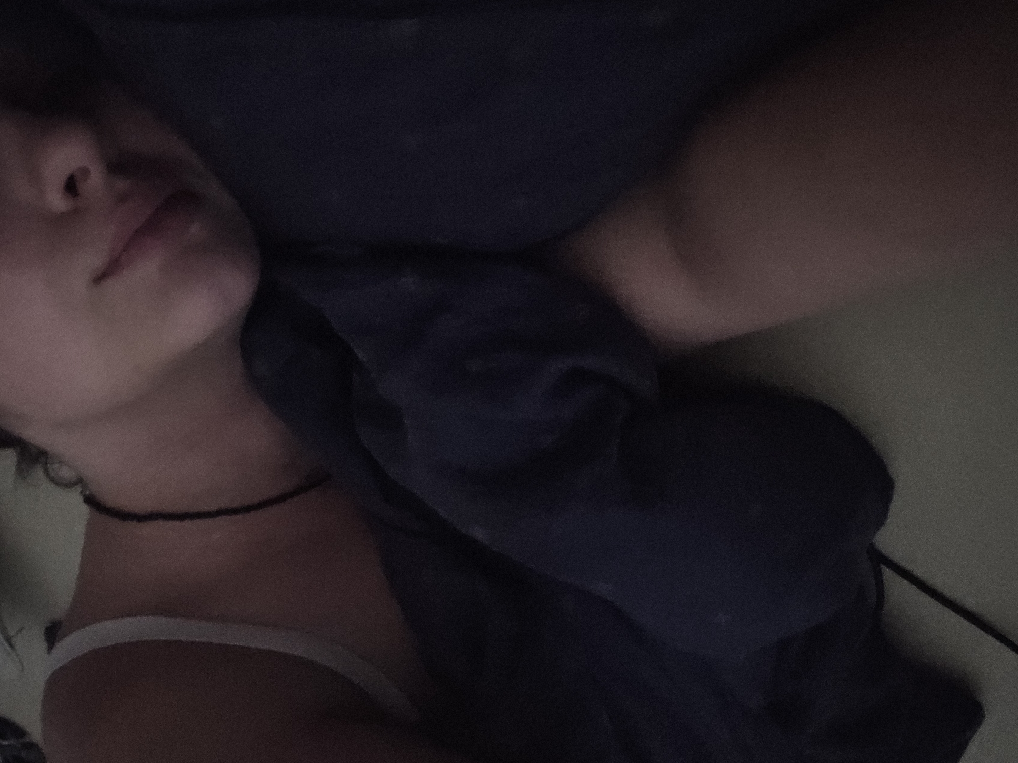 This is a close-up, overhead shot of a person lying down, partially covered by a dark blue blanket. The person's face is visible in the upper left quadrant, with their eyes closed and their head turned to the side. Their lips are slightly parted, and their skin tone is fair. A thin, dark choker necklace is visible around their neck, and they are wearing a white strap top or bra. The lighting is dim, suggesting it might be nighttime or a dimly lit room. The background is dark and indistinct, with the focus entirely on the person and the blanket. There are no visible text, weather cues, or specific location details beyond what can be inferred from the person's attire and the bedding. The overall impression is one of rest or sleep.