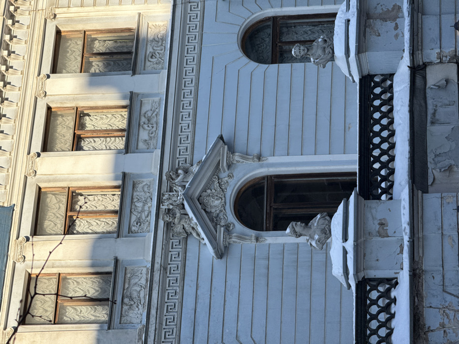 The image depicts a detailed view of the exterior facade of the House of the Former American Ambassador (O. P. Shtraus's apartment building) located at Bolshaya Konyushennaya Street, 13, Saint Petersburg, Russia.
The building presents a light blue, three-story classical-style facade, with the uppermost story partially cropped. The architecture is heavily ornamented with white stucco. The visible upper floor features three rectangular windows with dark wooden frames and light, patterned curtains. Below these windows are decorative bas-relief panels, appearing to depict figures or scenes. Below this, a prominent frieze with a repeating Greek key (meander) pattern runs horizontally across the facade.
The lower visible floor includes three arched windows with dark wooden frames and curtains. The central arched window is surmounted by an elaborate pediment-like stucco decoration. This decoration features a central cartouche with a human head, flanked by two winged cherubic figures (putti) holding a shield or crest. Below these windows is a balcony with a dark, ornate metal railing. Two classical-style female busts, carved from light-colored stone, are positioned on the stone pedestals that cap the balcony railing; both busts are partially covered with snow.
The scene is set in winter, with snow accumulated on ledges, the balcony railing, and the busts. Direct sunlight illuminates the upper-right portion of the facade, creating shadows on the left side. A bare tree branch is visible in front of the upper-left windows. The building's facade shows signs of age and wear, including areas of peeling paint or plaster on the lower walls and weathered stucco ornamentation. No people, actions, or interactions are visible.
