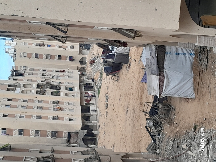 This image depicts a severely damaged urban landscape in Khan Yunis, Palestinian Territory, characterized by widespread destruction of multi-story residential buildings. The scene is viewed from an elevated perspective, looking down into a sandy, debris-strewn open area.
In the background and midground, several tall, light-colored apartment buildings show extensive structural damage, including numerous pockmarks on their facades, shattered or missing windows, and bent metal window grates. Sections of balconies appear damaged, with some fabric or tarps draped from them. Debris hangs from upper floors of one central building. The lower levels of these buildings feature arched doorways and damaged ground-floor sections.
The ground consists of unpaved sand interspersed with significant piles of concrete rubble, twisted metal, and other construction debris. In the midground, near the base of the central building, two figures are visible standing on the sandy ground amidst some rubble.
In the foreground and midground, several makeshift shelters are constructed from various materials. One prominent shelter in the lower right foreground is made of white tarpaulin with a blue "unicef" logo visible on its side, topped with dark, layered materials. Further into the midground, other less defined shelters, some dark in color and one possibly incorporating an inverted umbrella, are scattered across the sandy terrain. A metal frame, possibly from furniture, lies amidst rubble in the foreground. Items of clothing are visible hanging from windows and balconies on the damaged buildings. The foreground also includes a portion of another damaged building on the right, with arched elements and partially open windows featuring bent metal grates.