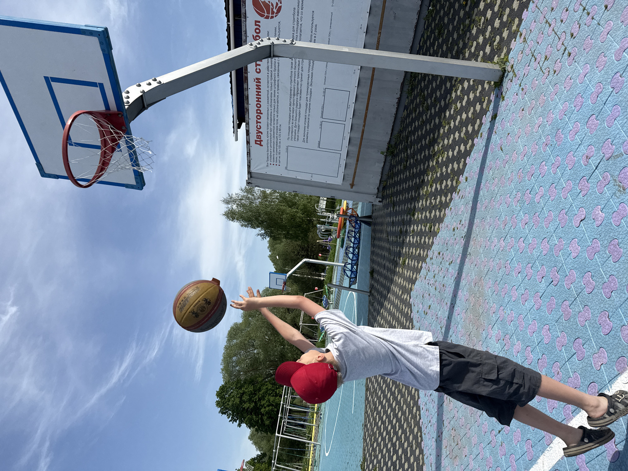 A young boy in a gray t-shirt, black shorts, and a red baseball cap is playing basketball on an outdoor court in Sin'kovo, Russia. He is extending his arms upwards toward the basketball, which is in mid-air between him and the basketball hoop. The court has a blue and purple tile pattern with black tiles on the sides. The basketball hoop and backboard are to the left. Behind the court are trees and in the background a white building with signage in Russian.