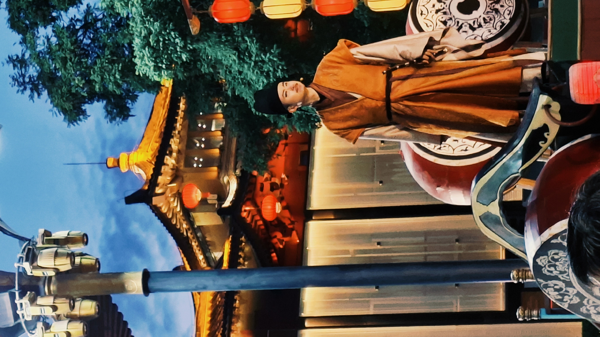 The image depicts a man dressed in traditional Chinese clothing standing on a decorated platform, likely part of a performance or cultural display. He is wearing a dark cap and a golden-brown robe with a black belt. Behind him is a building with traditional Chinese architecture, adorned with red lanterns. Lush green foliage is visible near the building. The foreground includes a metal pole and decorative objects. The location is likely in Yanta District, China, given the traditional Chinese architectural style and cultural attire.