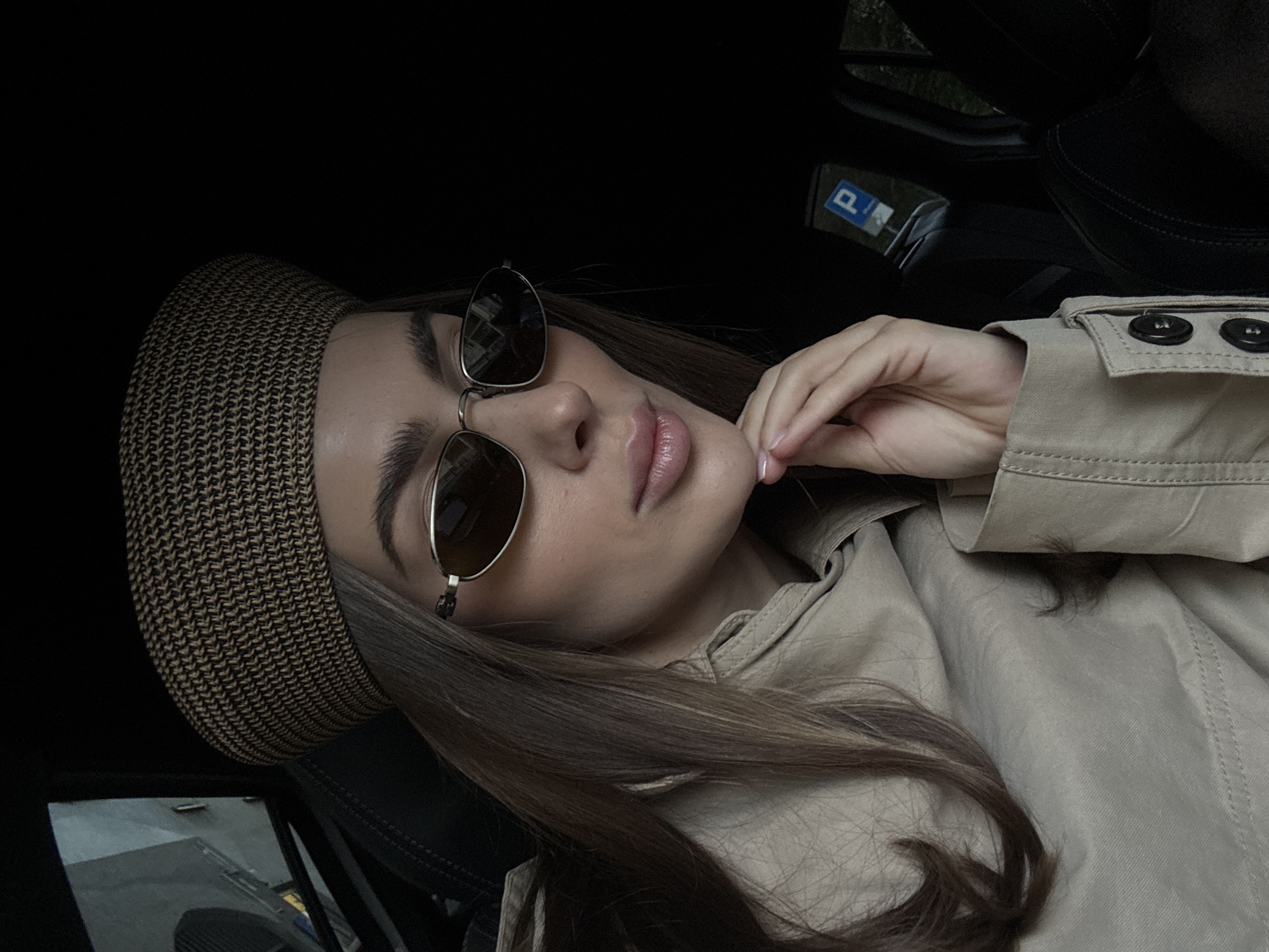 A woman is in a car wearing a brown beret, brown sunglasses, and a tan trench coat. Her right hand is by her face. The car appears to be parked as there is a "P" parking sign visible. The location may be Warsaw, Poland, based on the "P" sign for "Parkowanie", which is parking in Polish.