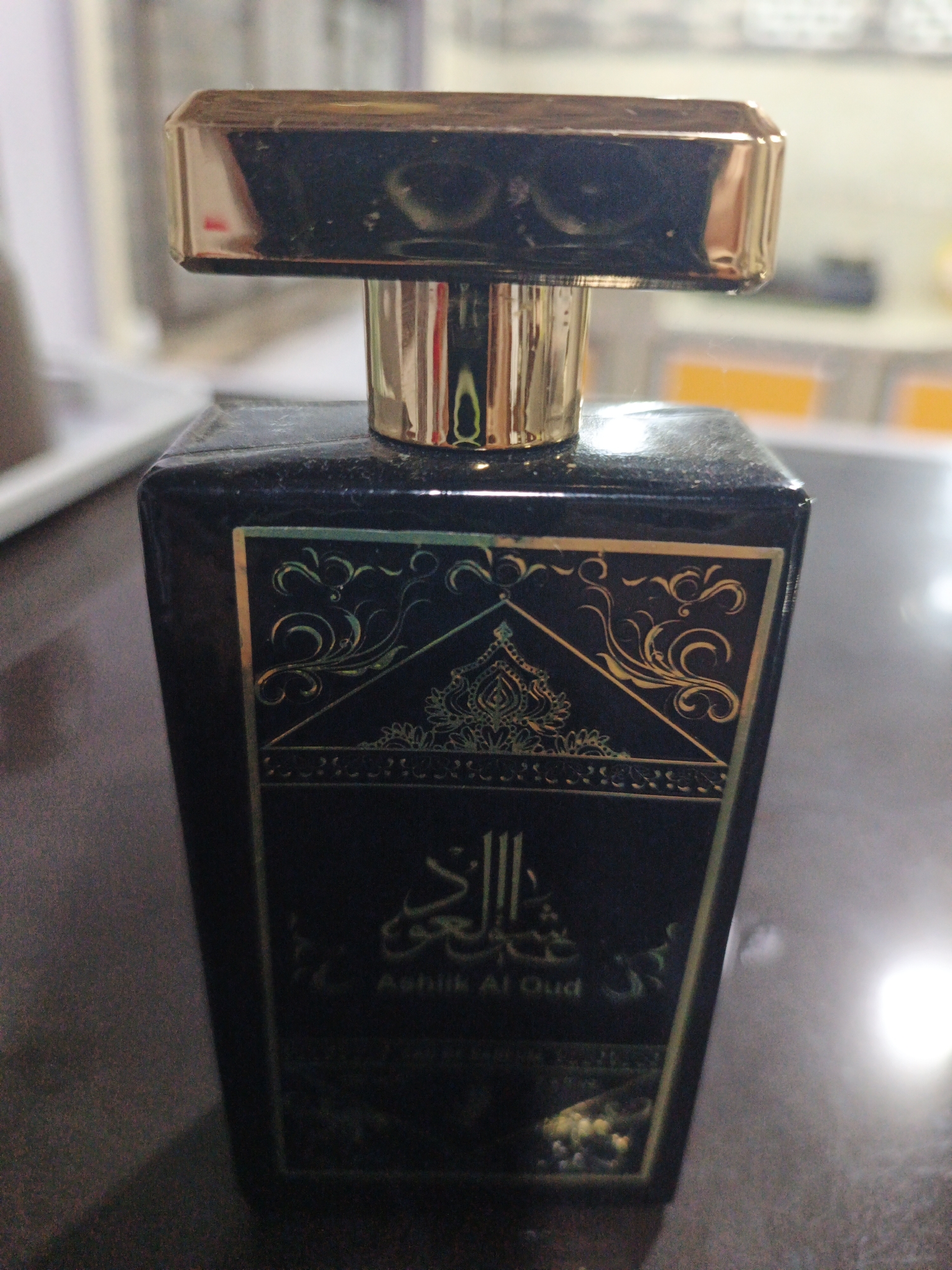 A black rectangular bottle of perfume with gold accents sits on a dark surface. The bottle has intricate gold patterns on its front. Arabic script and the words "Ashik Al Oud" are visible on the label. The background is blurred, suggesting an indoor setting. The location is stated as Kelapapati, Indonesia.
