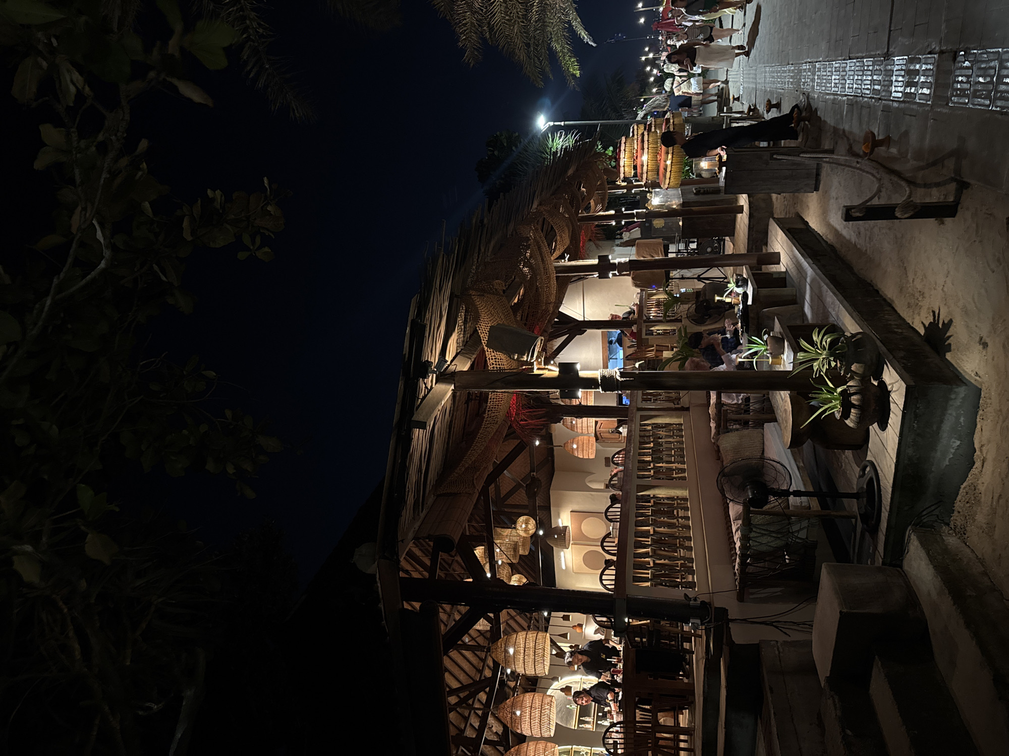 An outdoor restaurant in Kuta, Indonesia, is captured at night. The establishment features a thatched roof supported by wooden beams, adorned with fishing nets and large woven light fixtures. Guests are seated at wooden tables and chairs, some being served by staff. A fan is visible on a wooden platform near seating. In the background, a brightly lit walkway lines the beach, with palm trees and a streetlamp casting a glow. Pedestrians, casually dressed, stroll along the walkway. The scene is lit by ambient lights from the restaurant and the walkway, creating a warm and inviting atmosphere against the dark night sky.