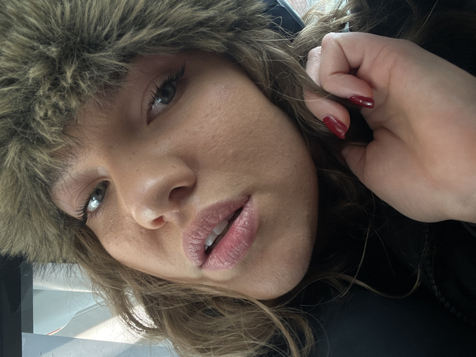 A close-up selfie depicts a female individual looking directly at the camera, head slightly tilted. She is wearing a brown, furry winter hat, possibly an ushanka style, which covers most of her hair. Her wavy, light brown hair is visible beneath the hat. She has light-colored eyes and her lips are slightly parted, revealing a sliver of her upper teeth. Her right hand, adorned with red nail polish, is positioned near her chin and neck. She wears dark-colored clothing, likely a jacket, visible around her collarbone. The background is indistinct and out-of-focus, suggesting an interior setting with some reflections. The scene is set in Moscow, Russia.