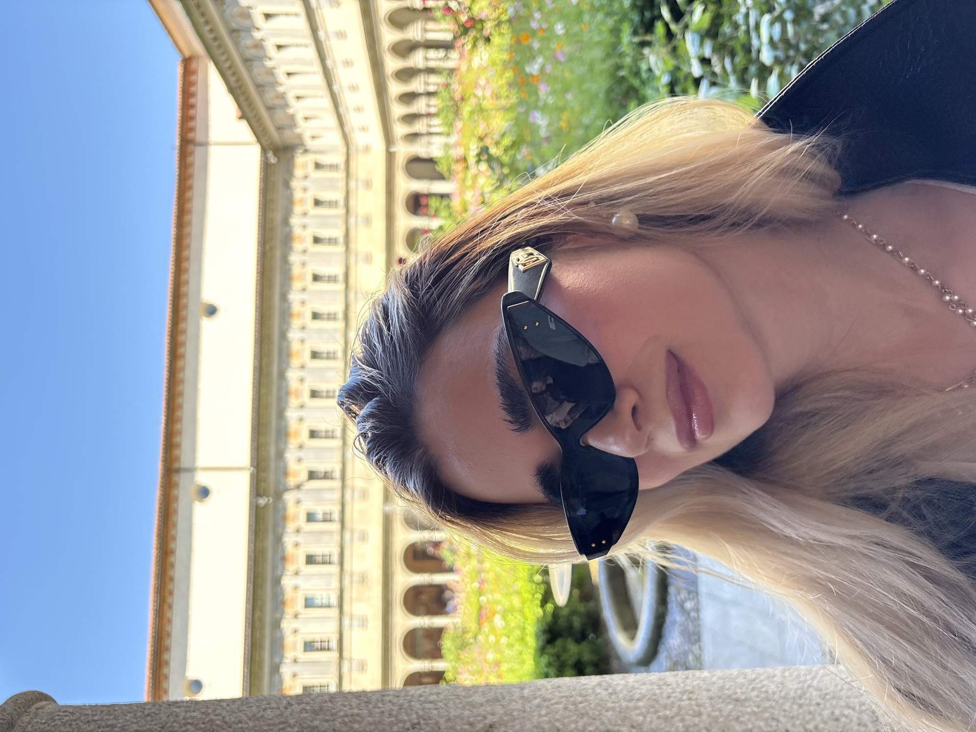 A woman wearing dark sunglasses poses outdoors with a building in the background, possibly in Milan, Italy. The building has arched windows and detailed stonework. To the right of the woman, there are flowers and greenery. The sky is clear and blue. The woman has blonde hair, and is wearing a pearl earring and necklace.