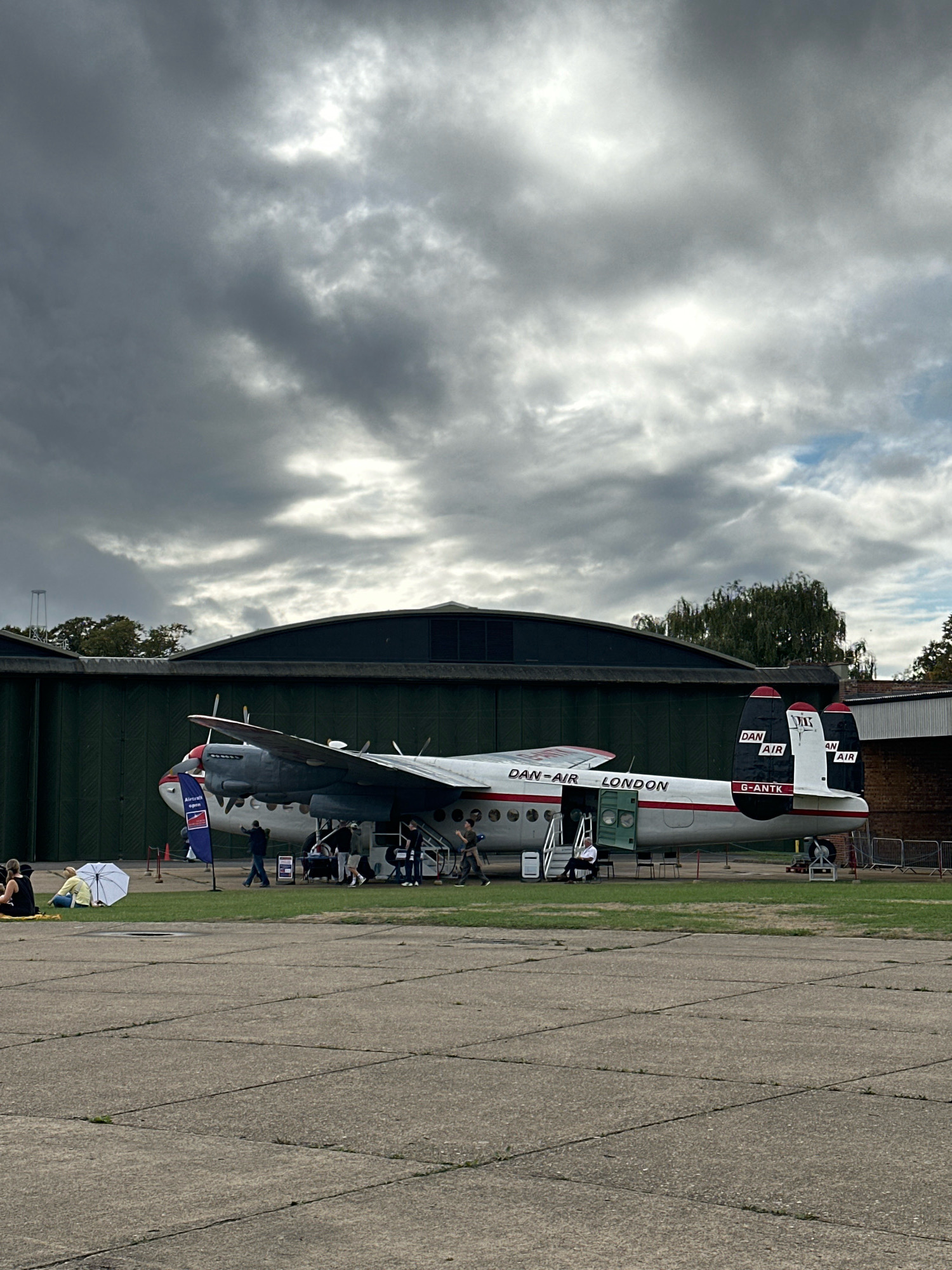An outdoor, eye-level, medium-shot captures a historical Dan-Air London aircraft, identified by its registration G-ANTK, on an overcast day. The plane is positioned on a tarmac in front of a dark green, curved hangar. The aircraft is white with red accents and features dual propellers. Its main cabin door is open, revealing stairs leading inside, with several people visible around the aircraft, some entering or exiting. In the foreground, a white umbrella is deployed next to two individuals sitting on the ground. The sky is heavily clouded, with patches of bright light suggesting sunlight breaking through. The scene appears to be at an aviation museum or heritage site, possibly within or near the city of Cambridge, United Kingdom, given the presence of historical aircraft and aviation-related infrastructure.