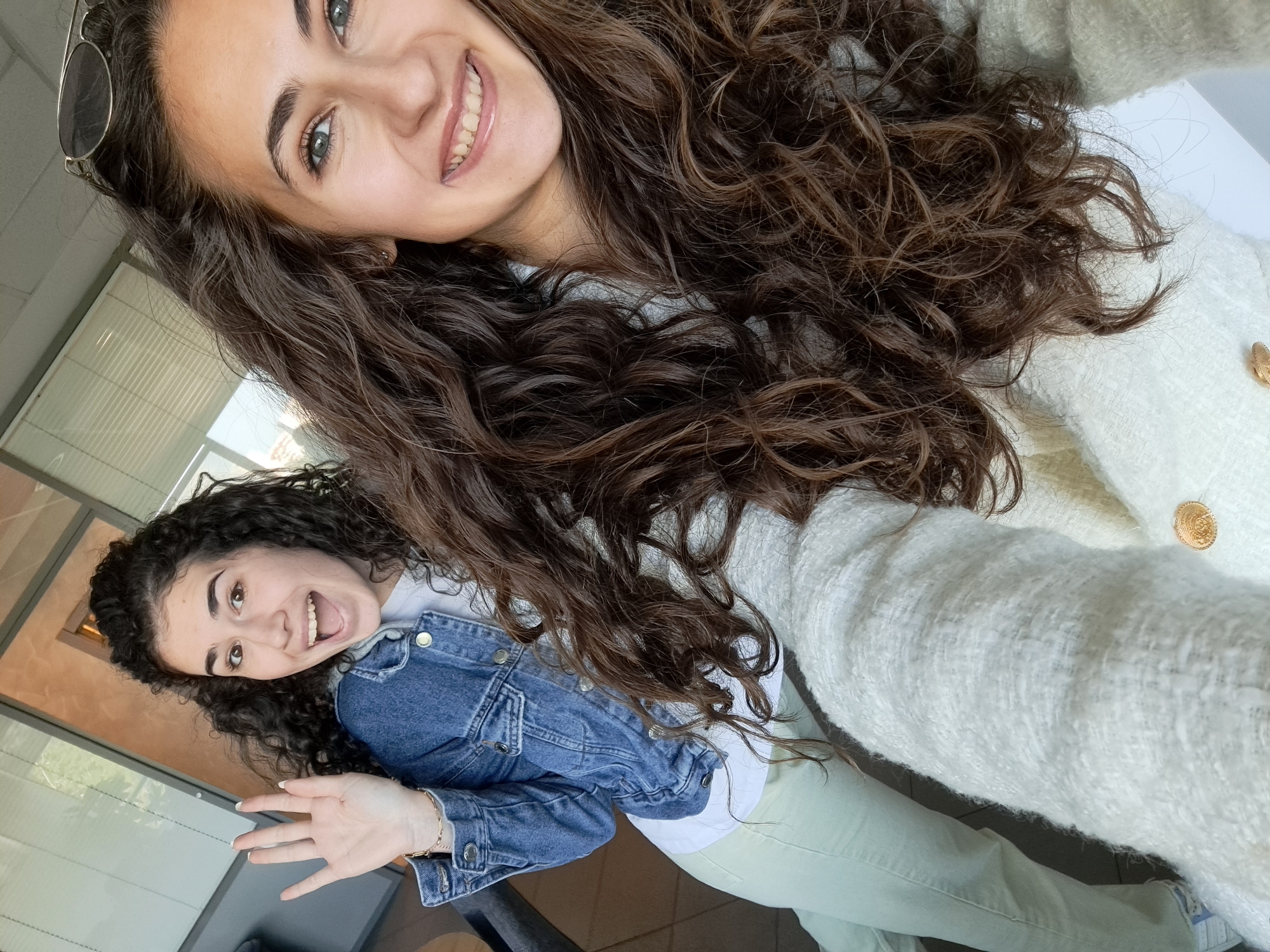 Two women are posing for a selfie in an indoor environment. The woman in the upper portion of the frame has long, dark, curly hair, wearing a light-colored tweed sweater with gold buttons and sunglasses resting on her head. She is smiling broadly. The second woman is located in the lower left area of the frame, wearing a denim jacket over a white top. She has curly, dark hair pulled back from her face and is sticking her tongue out while waving her hand. They are both positioned near a window with blinds. The background indicates a room with office or institutional features, situated in the city of Unknown, Unknown.