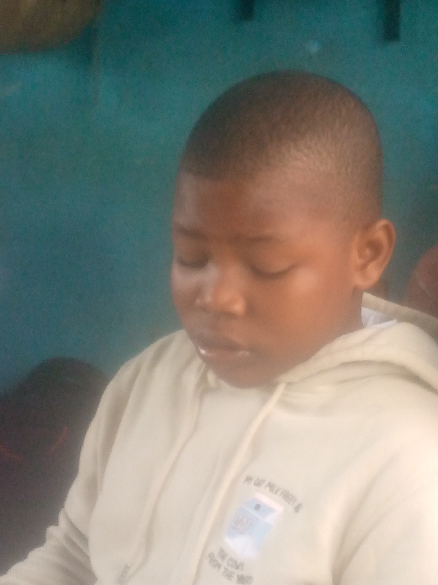 A young boy with short, dark hair is seen indoors, wearing a light-colored hoodie. His eyes are closed, and he appears to be looking down. The background is a teal-colored wall. The location, Ikorodu, Nigeria, cannot be determined from the image alone.