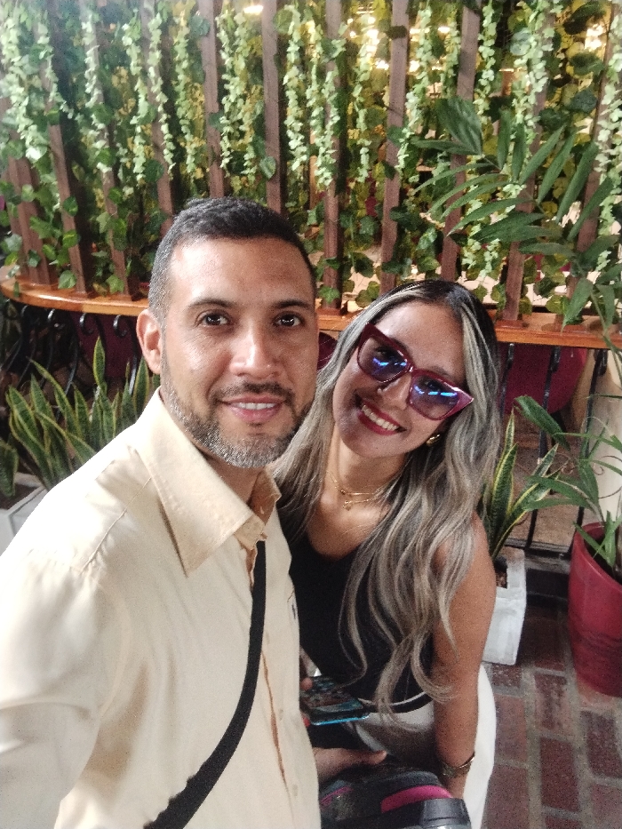 A selfie captures a smiling man and woman in an outdoor setting. The man, with short dark hair and a salt-and-pepper beard, wears a light-colored shirt with a black strap across his chest. The woman, with long silver hair and striking pink sunglasses, wears a black top. Lush greenery forms the backdrop with various potted plants visible. The ground appears to be brick. Both individuals are looking directly at the camera, conveying a joyful mood.