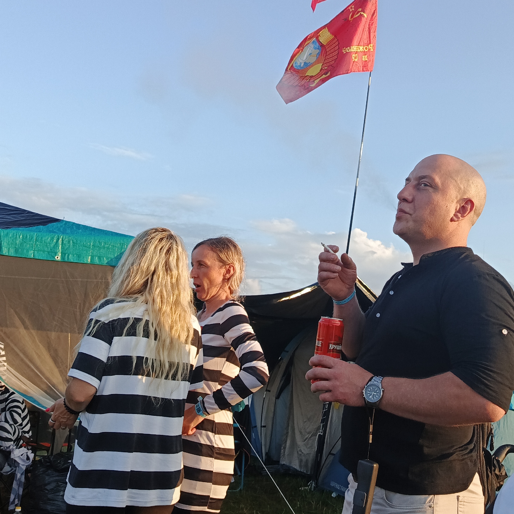 The image depicts an outdoor scene, possibly a campground or festival, with tents visible in the background. Three individuals are the primary subjects. On the left, a person with long blonde hair, wearing a black and white striped shirt, faces away from the camera. To their right, a woman with short blonde hair, also wearing a black and white striped shirt, is engaged in conversation, with her mouth open as if speaking. On the right side of the image, a bald man, wearing a black shirt and light-colored pants, holds a red can in his right hand and a cigarette in his left hand, looking upwards. He is wearing a wristwatch on his left wrist. A red flag with a Soviet emblem is visible flying in the sky above the man's head. The overall lighting suggests it is daytime, likely late afternoon or early evening, with a clear sky and some scattered clouds. The location is identified as Lagunovo, Russia.