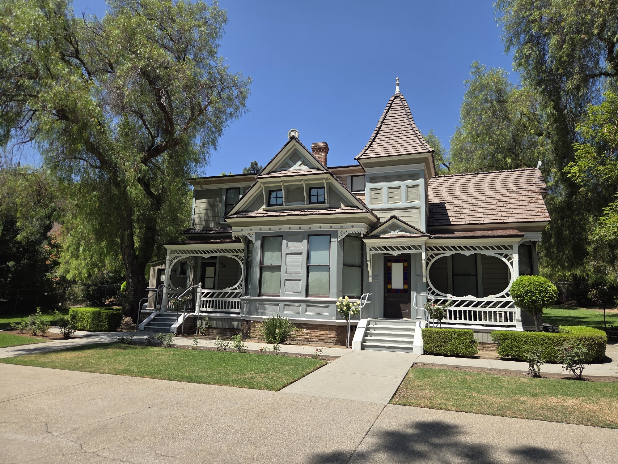 This image captures a well-maintained, two-story Victorian-style house located in Glendale, USA. The house features a prominent turret with a conical roof, numerous windows of various shapes and sizes, and intricate decorative trim. The exterior is painted in muted tones of green and gray, with a brown shingled roof. A wrap-around porch with white railings and ornate spindles enhances the architectural charm. The house is surrounded by a lush green lawn, manicured hedges, and mature trees, suggesting a suburban or park-like setting. A concrete sidewalk leads to the front entrance, and the street in the foreground is also paved. The sky above is a clear, bright blue, indicating a sunny day. No people or animals are visible in the image.