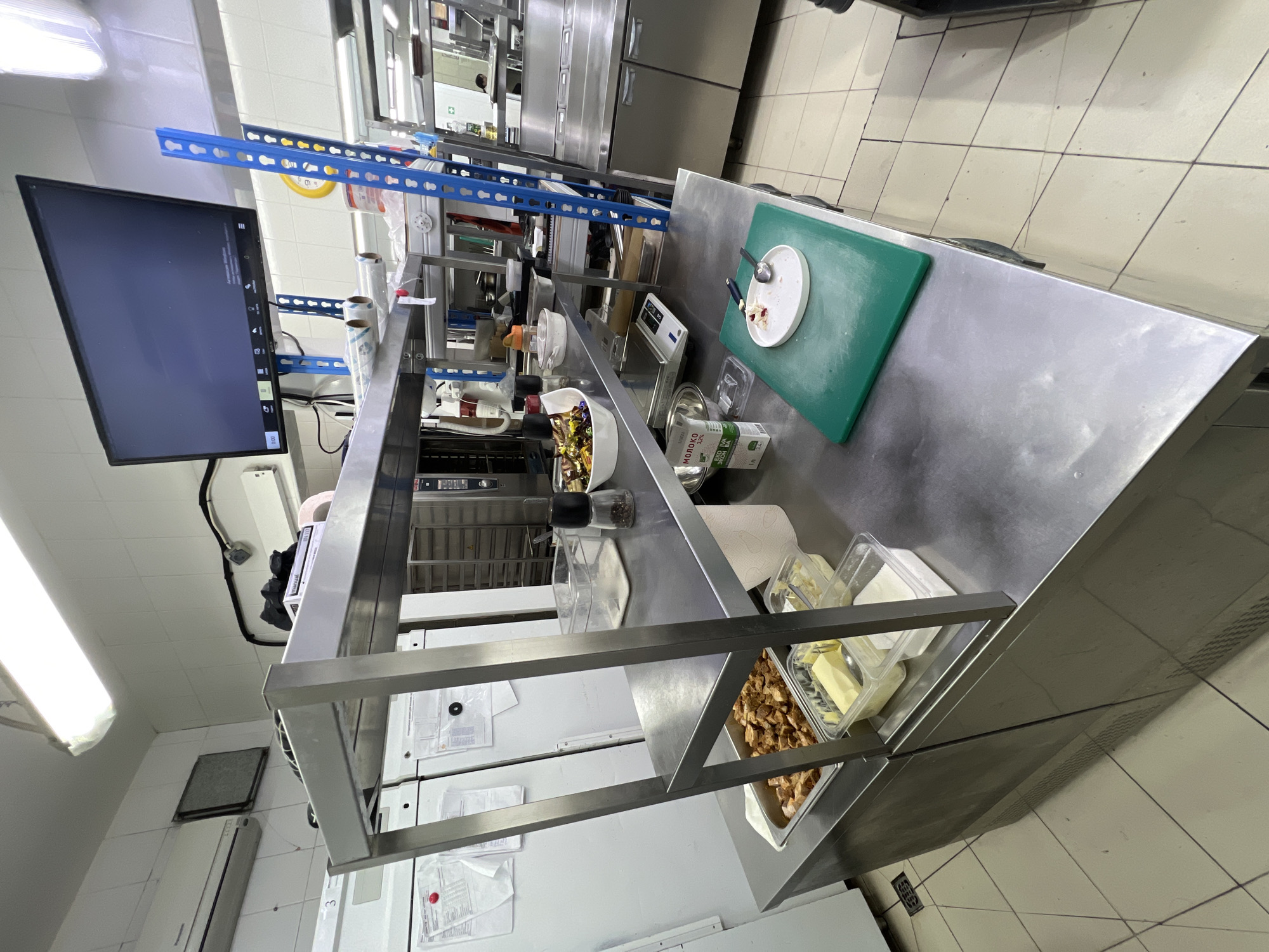 This is a wide-angle, eye-level photograph of a commercial kitchen environment. The scene is brightly lit, likely by fluorescent overhead lights, and captured in daylight. The camera is positioned at an angle that provides a clear view of a stainless steel preparation counter, shelves laden with various kitchen supplies, and a portion of industrial kitchen equipment.
Dominating the foreground is the stainless steel preparation counter. On it, a green cutting board is visible with a white plate containing a small amount of food residue and two spoons. Adjacent to this is a bowl filled with a mixture of food items, possibly a salad, and a pepper grinder. Further to the left on the counter are several clear plastic containers, one holding what appears to be butter, and another with white granulated sugar. A metal scale is also present on the counter.
Above the counter, a two-tiered stainless steel shelf unit holds a variety of items. The lower shelf contains a stainless steel tray filled with diced meat, likely chicken or pork, and more plastic containers. The upper shelf has additional containers, rolls of what appears to be plastic wrap or similar material, and a black object that could be a book or manual.
In the background, a large flat-screen monitor is mounted on a blue metal shelving unit. The monitor displays a dark screen with some text visible, possibly a control panel or menu. The blue shelving unit is tall and industrial, holding various items. Behind the shelves, industrial kitchen appliances are visible, including what look like ovens or proofers. To the left of the preparation counter, large white appliances, possibly refrigerators or freezers, are partially visible. The walls of the kitchen are tiled in white, and the floor is tiled in a light beige color with dark grout lines.
There are no people clearly visible in the frame. The overall impression is one of an organized, active food preparation space.
The location is identified as Plast univska, Russia.