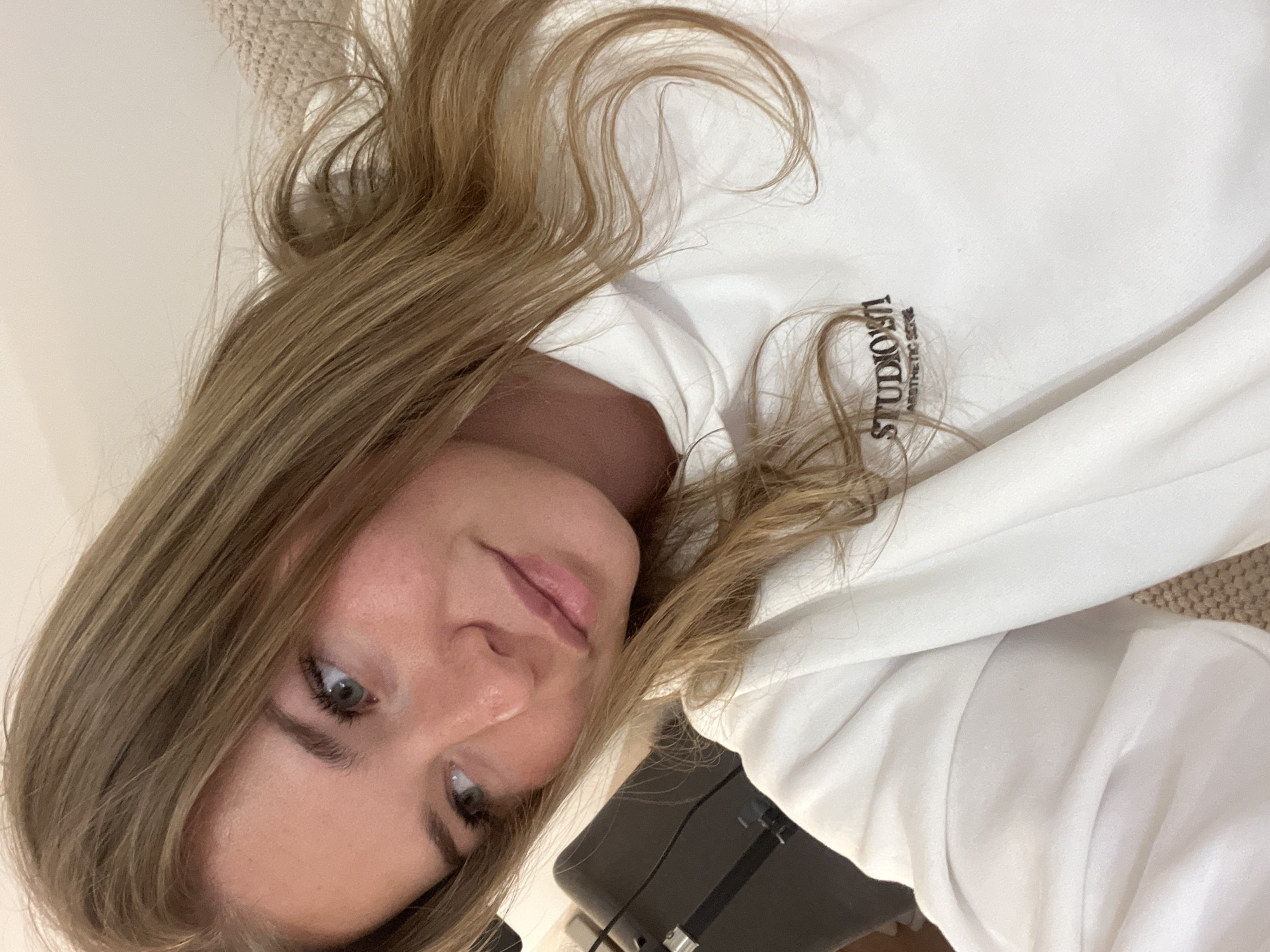 A medium close-up shows a fair-skinned young woman with light brown hair wearing a white sweatshirt with the text "STUDIOFI" printed in black. The camera is positioned above and to the right, capturing her face at an angle. She looks toward the camera with a slight smile. Her eyes are light in color and her hair is long and styled. The background is blurred, showing a textured surface, likely a beige carpet or upholstered furniture. A black object is visible in the lower part of the image, partially obscured. There is no evidence in the image to suggest the location is Budva, Montenegro.
