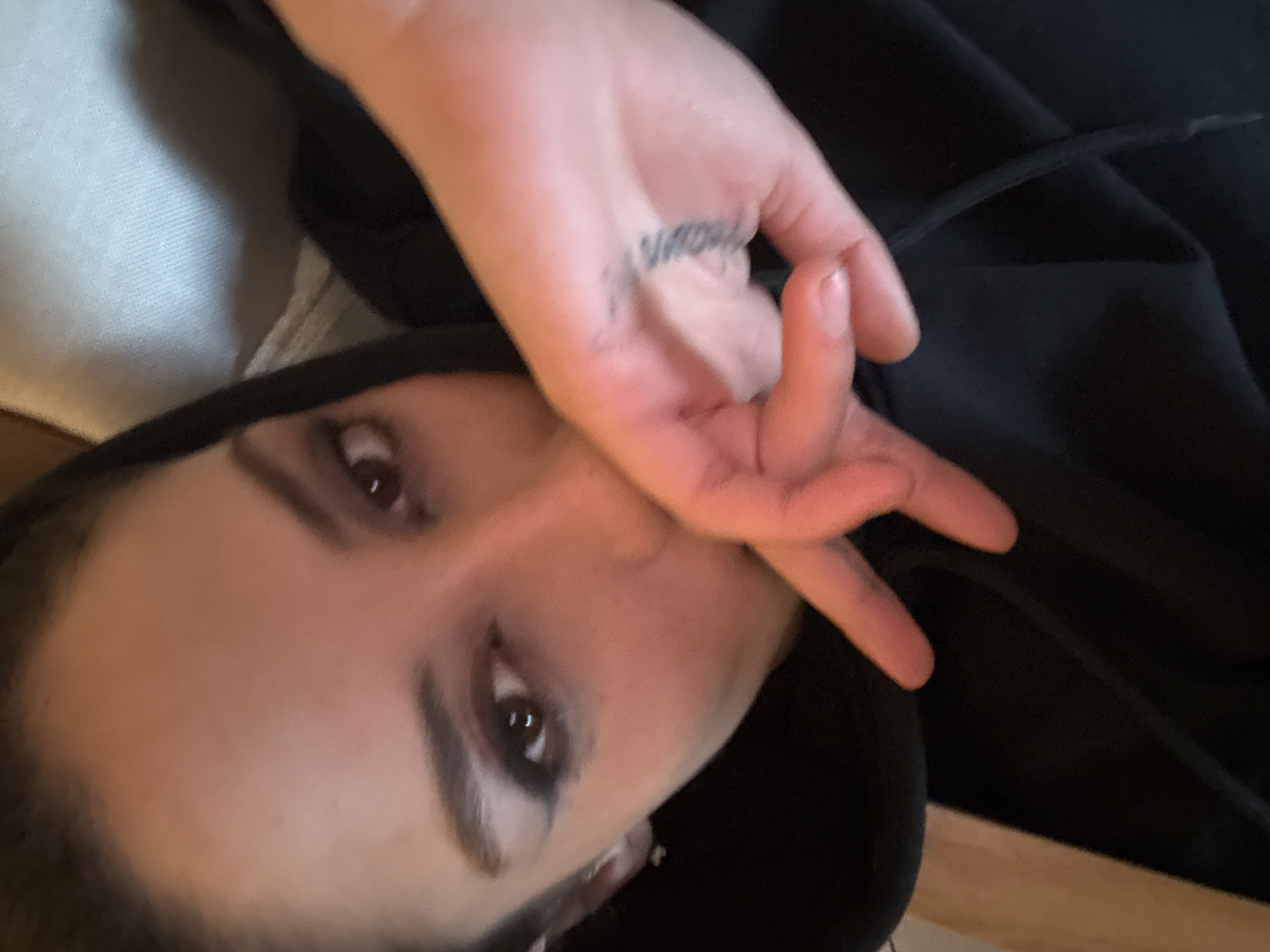 A close-up, rotated view of a person with fair skin and dark hair, wearing a black hooded garment, is shown. The person's face is angled, with dark eyes and prominent eyebrows. The left hand is raised and positioned in front of the face, displaying a small tattoo on the side of the hand. The background includes a light-colored surface. There is no indication of Buenos Aires, Argentina.
