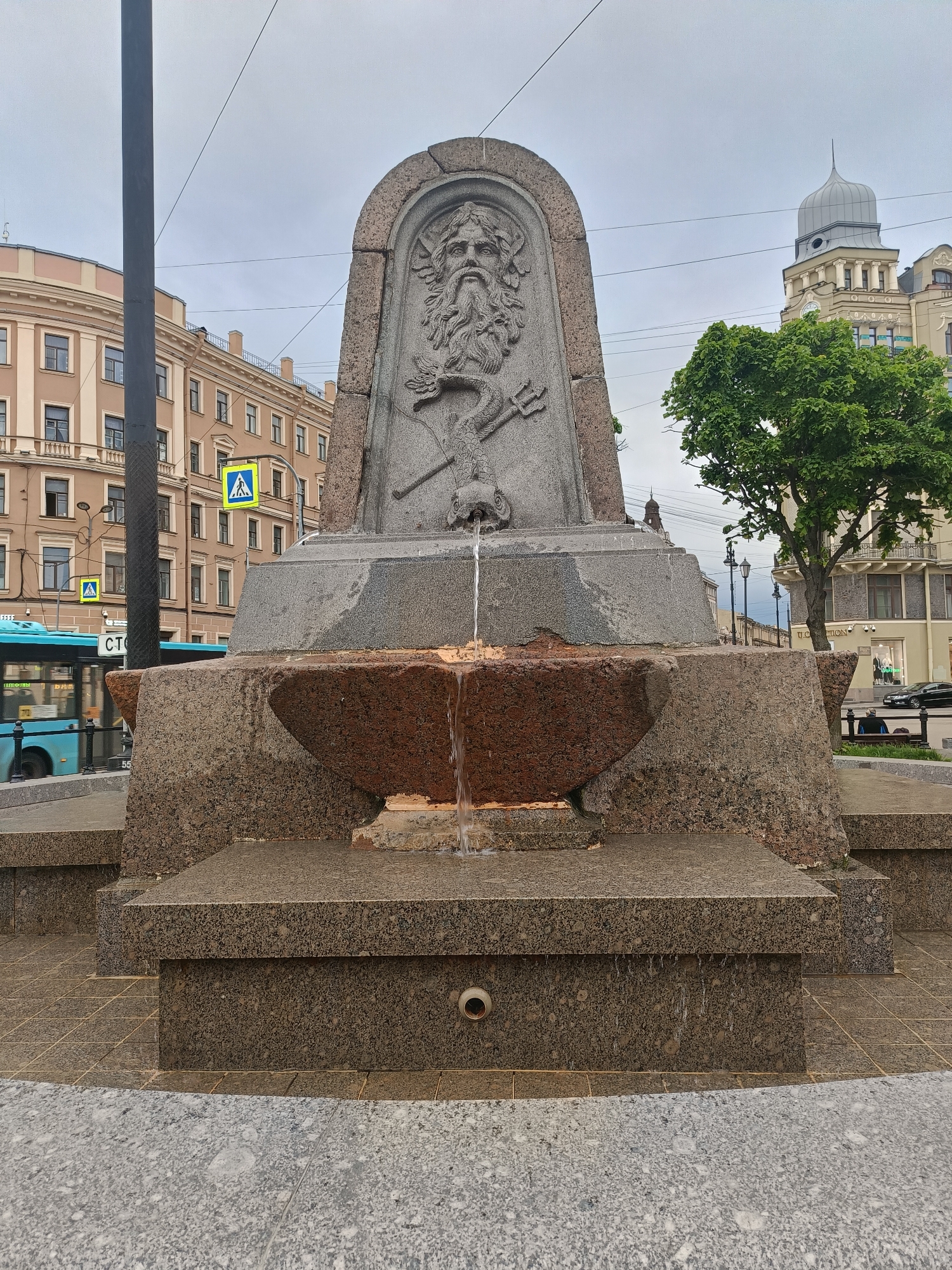 The image shows a fountain featuring a relief of Neptune with a trident in Saint Petersburg, Russia. The fountain is constructed of granite. The relief of Neptune is above a basin from which water flows into a lower level. Buildings are visible in the background on a cloudy day. A blue bus is partially visible to the left. A tree is visible to the right.