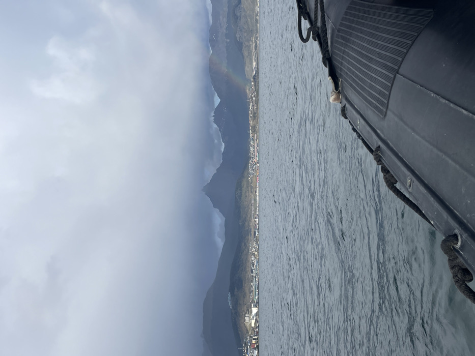 A view from a boat onto a coastal scene. Mountains rise from the edge of a town under a cloudy sky. A rainbow arches over the mountains. The water is textured, with ripples and reflections. The side of a dark boat with ropes and detailing is visible on the right. The overall scene is moody and scenic.